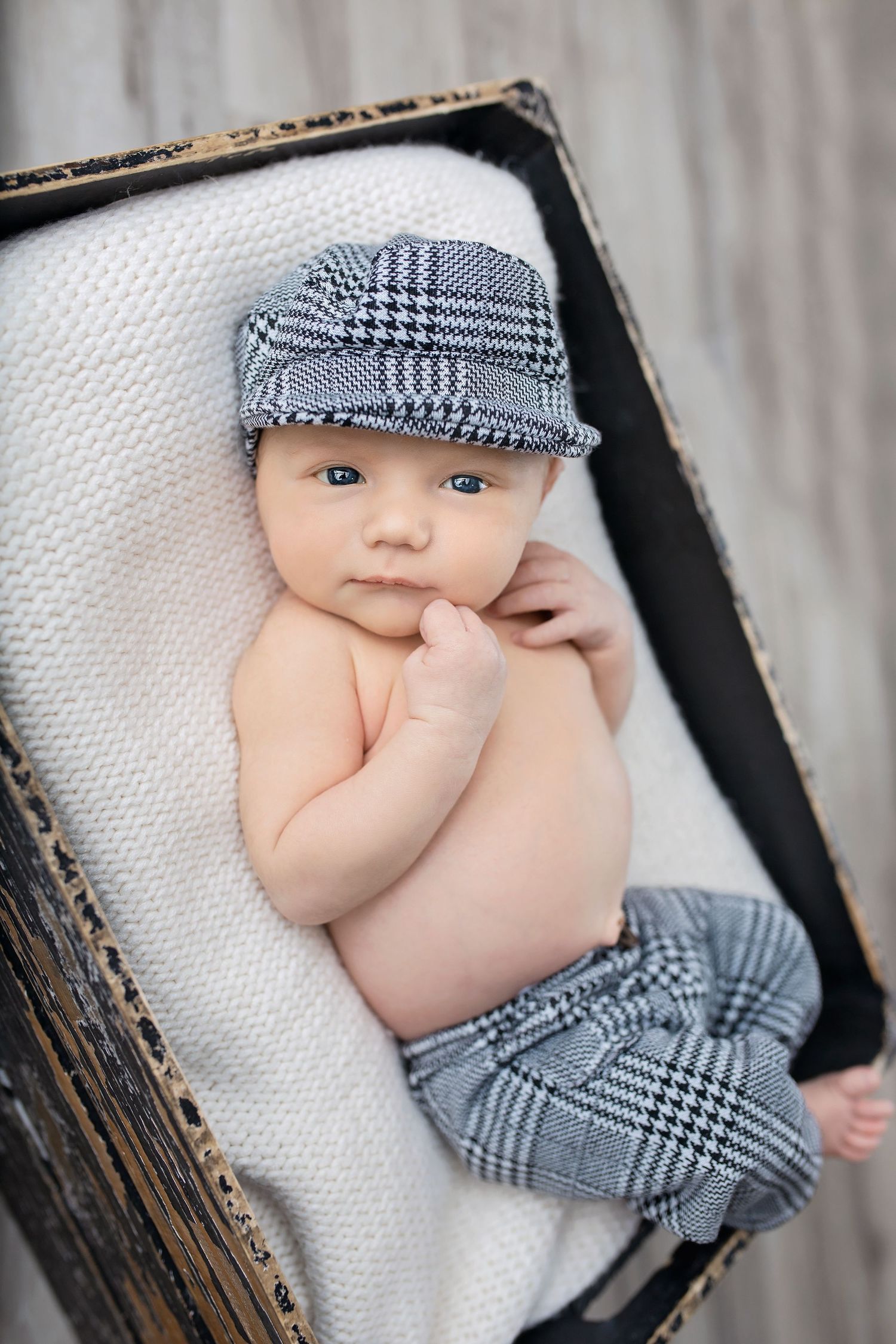 Alert newborn baby wearing plaid newsboy cap and matching pants lying in a dark wooden box.