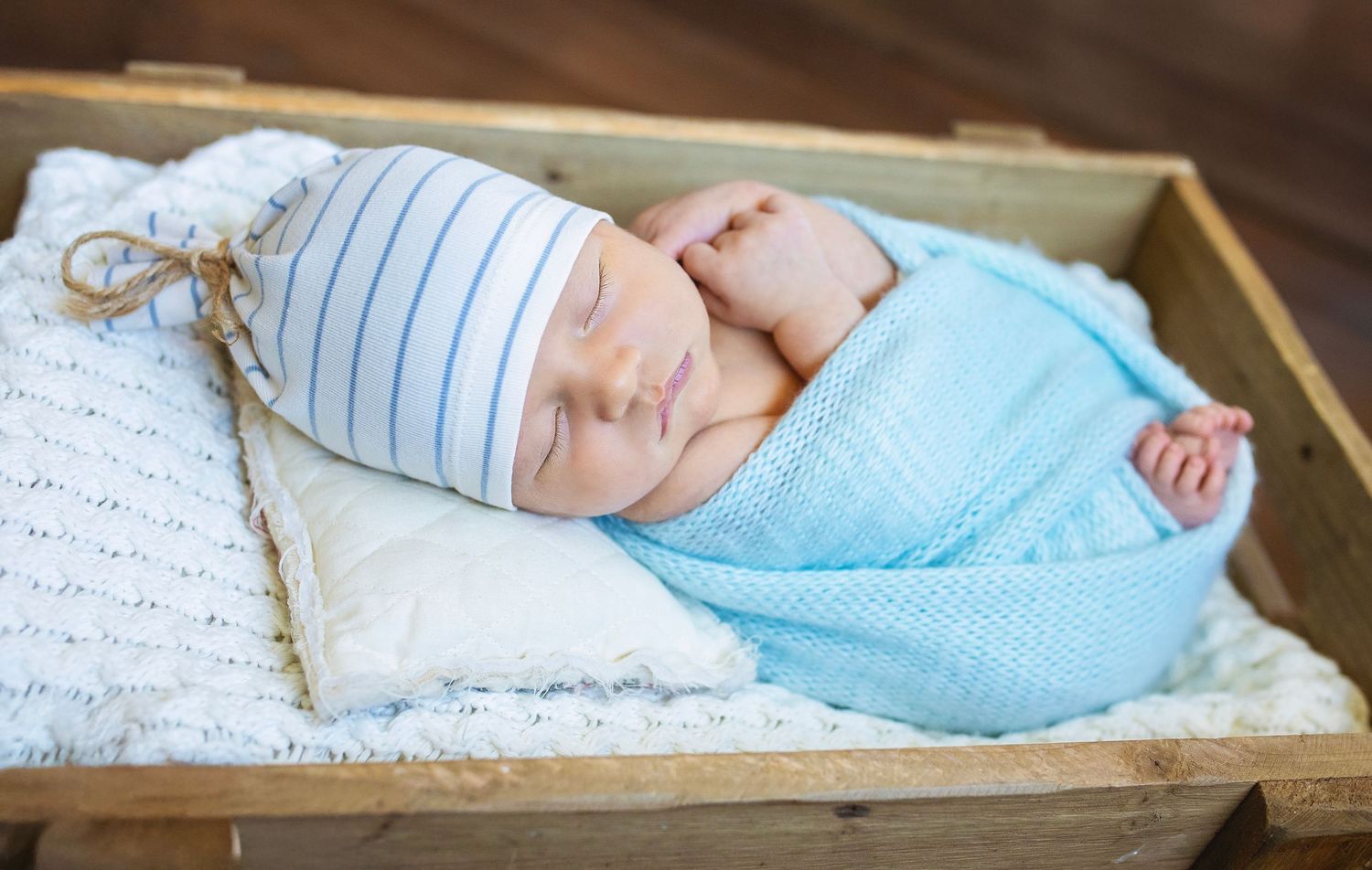 Peaceful newborn baby sleeping swaddled in blue wrap with striped hat in a wooden crate.