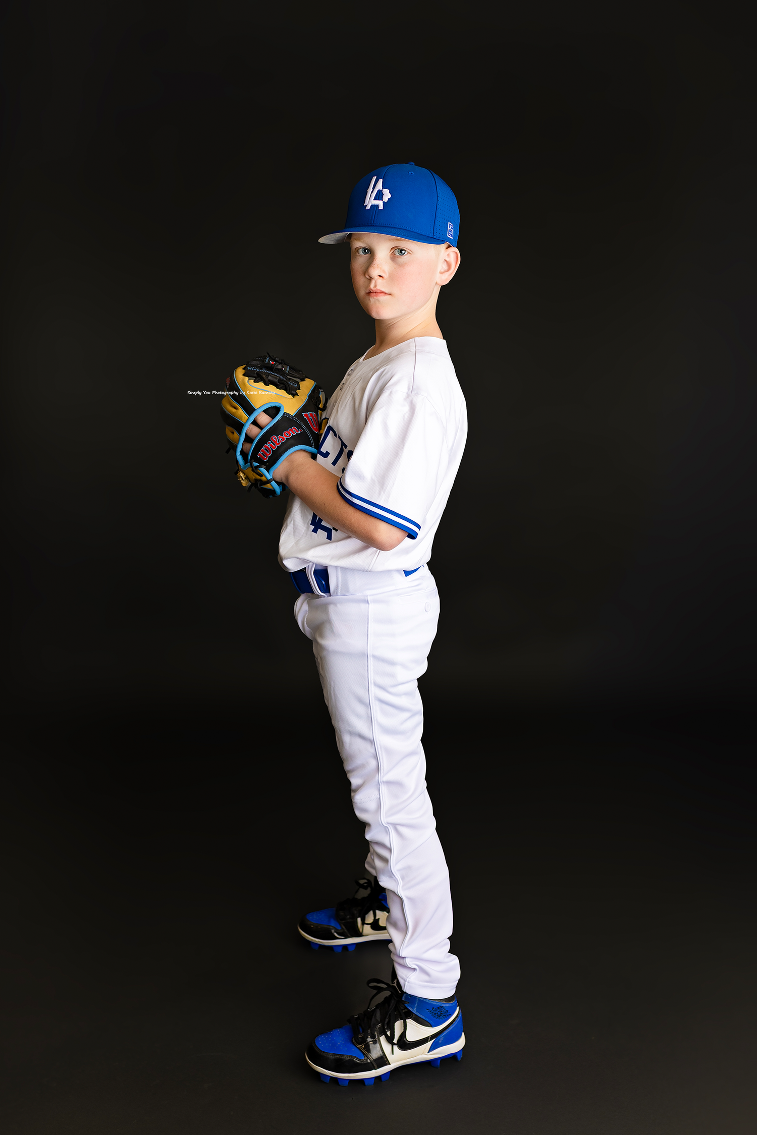 Young boy in white baseball uniform and blue cap poses with glove on black background.