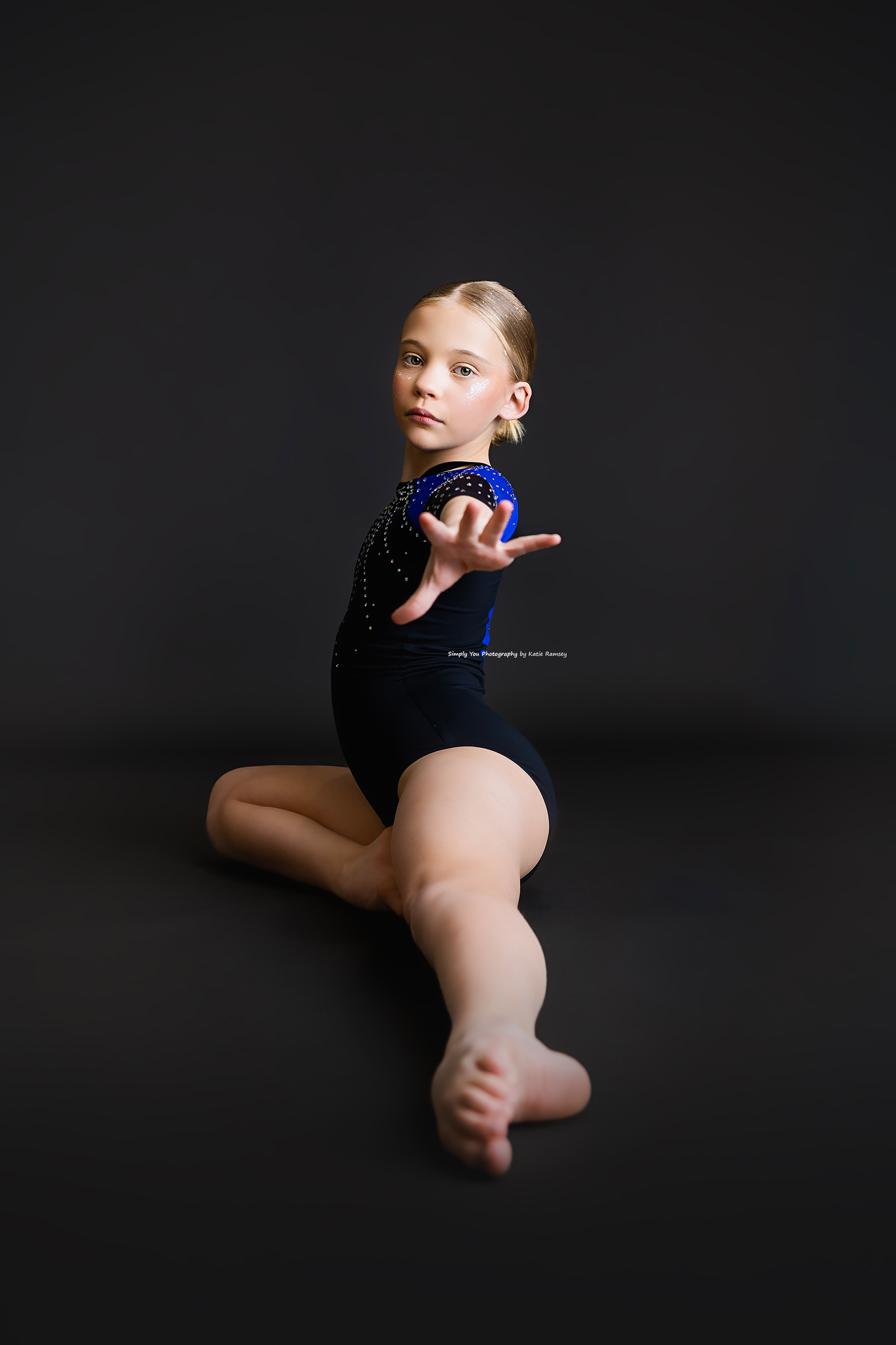 Young female gymnast in black leotard poses gracefully on floor during professional studio photo session.