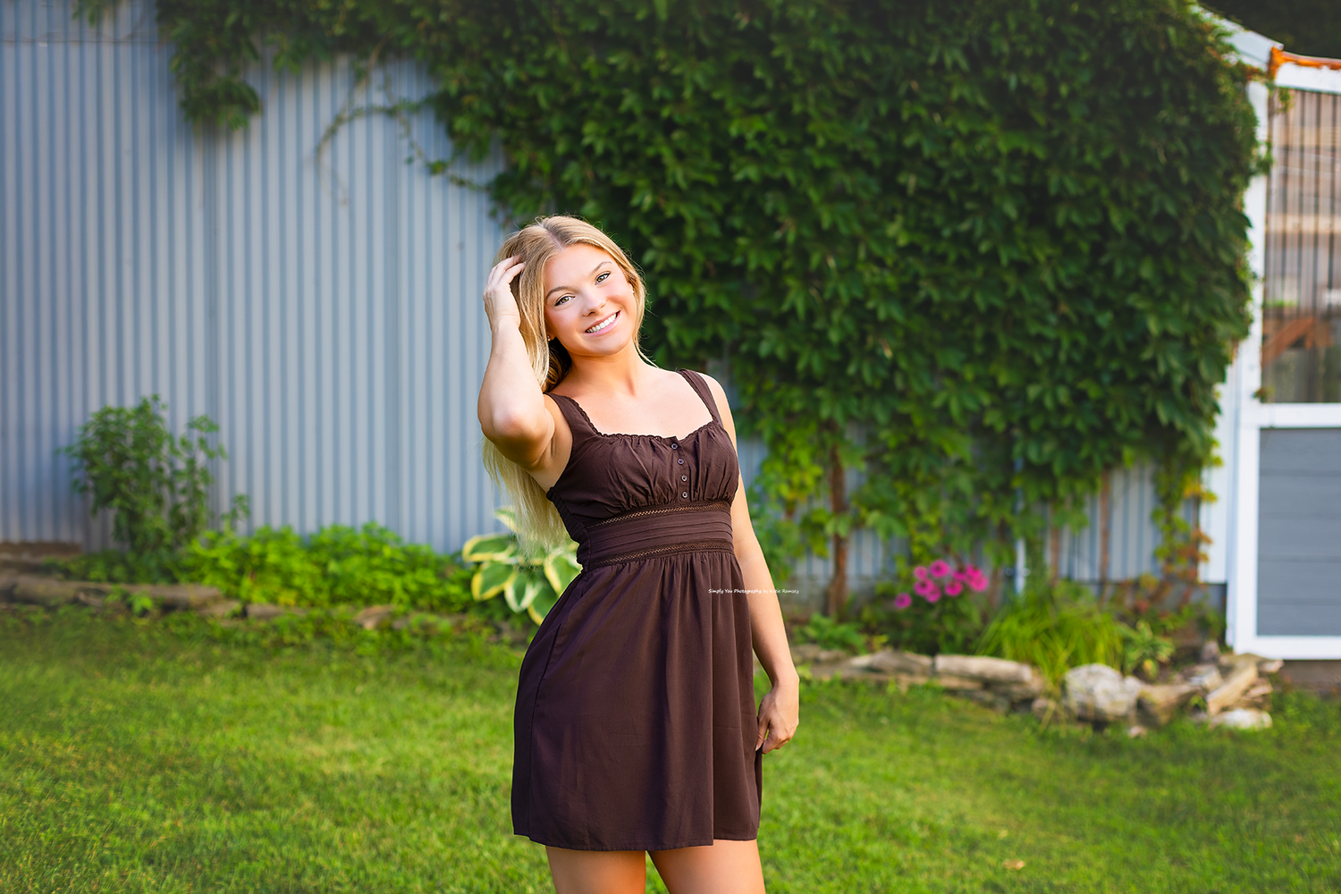 Smiling blonde woman in brown sundress posing outdoors on green lawn with ivy-covered building in background.