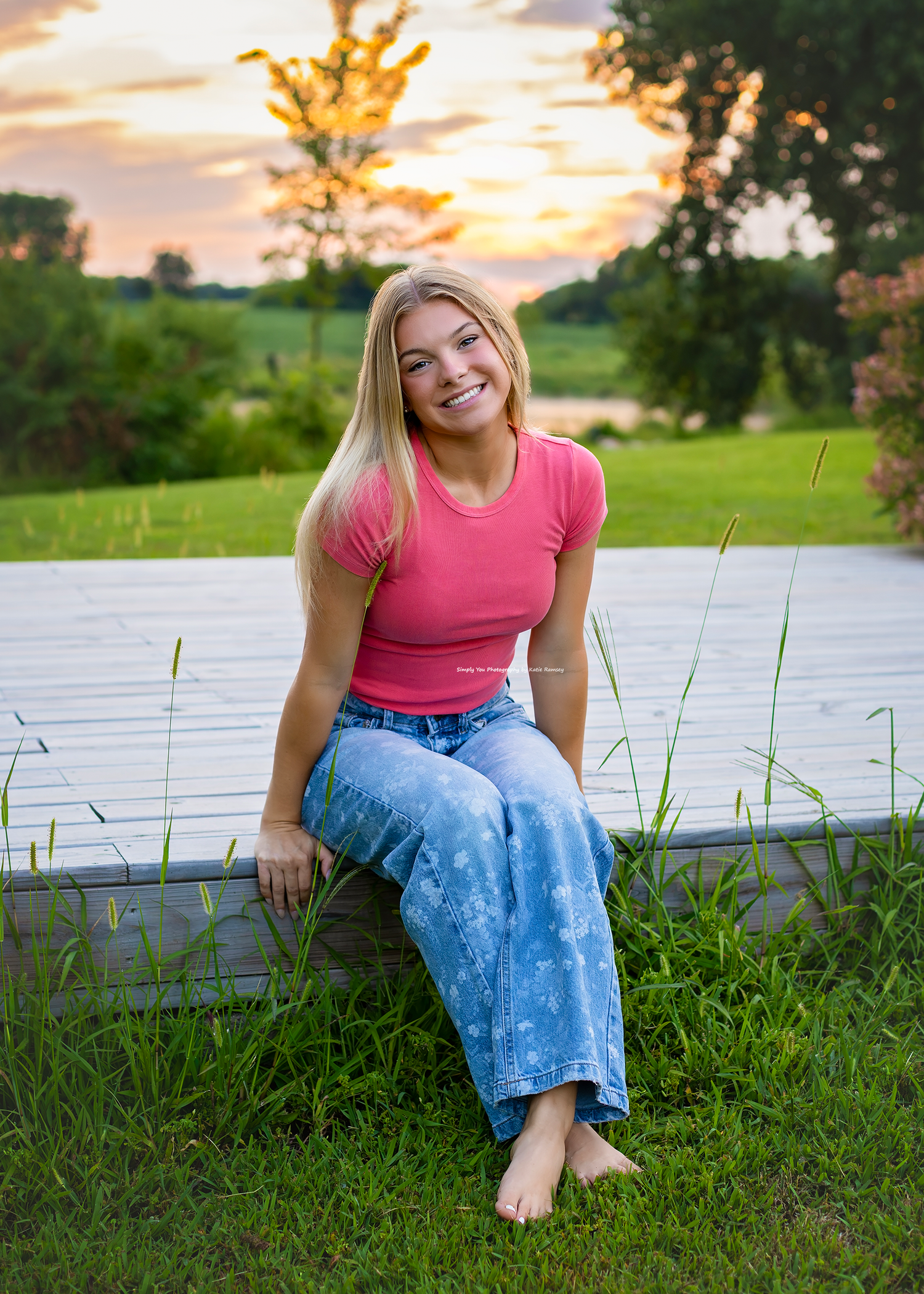 Smiling blonde teen girl in pink top and jeans sits barefoot on wooden deck at golden hour sunset.