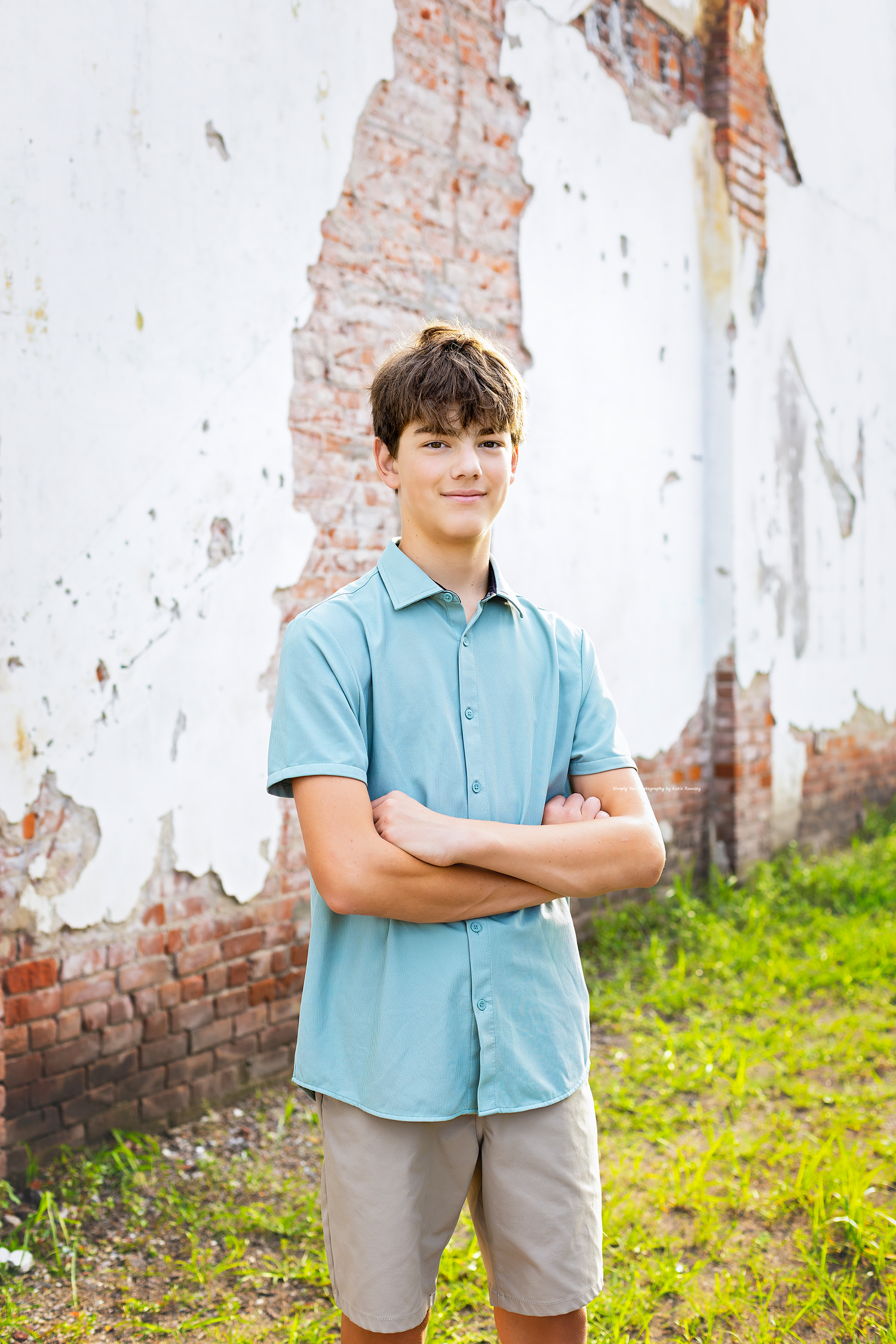 Teen boy in light blue shirt and khaki shorts stands confidently with arms crossed against a weathered brick wall.
