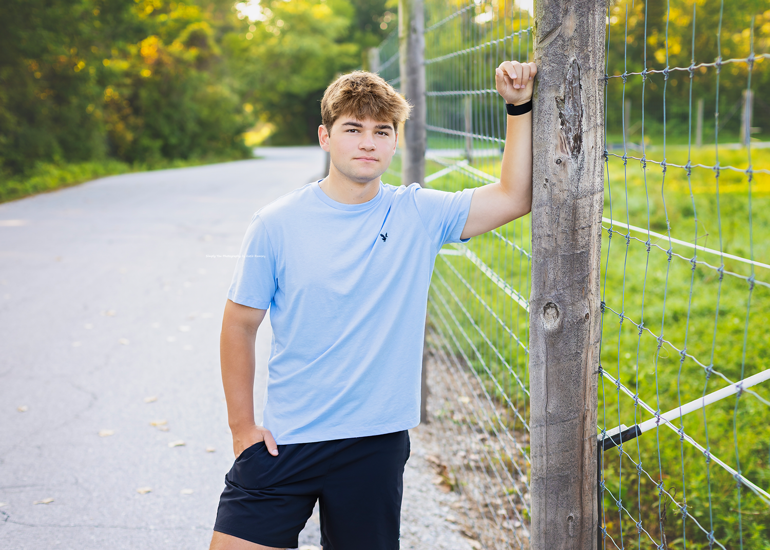 A teenage boy in a light blue shirt leans against a wooden fence post on a tree-lined country road.