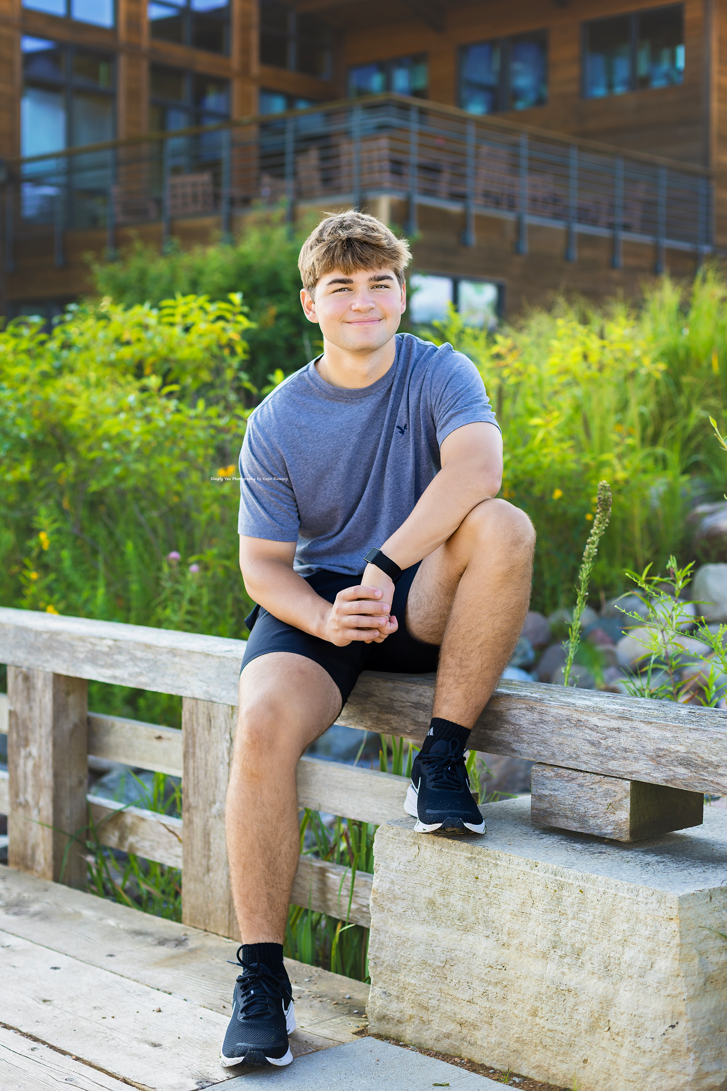 Young athletic teen sits on wooden bench outdoors in gray shirt and black shorts, smiling at camera.