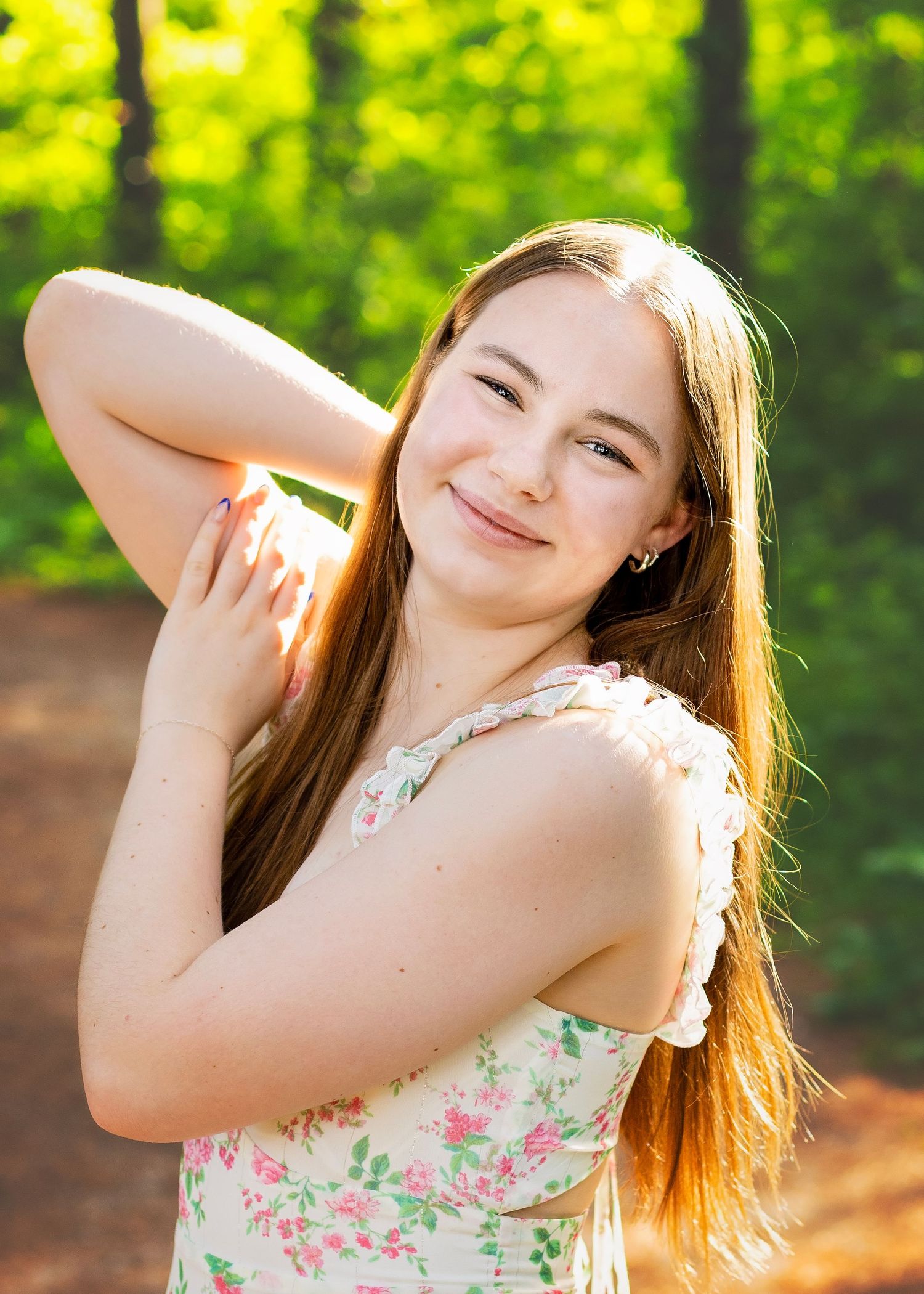 Smiling teenage girl in floral dress posing outdoors in sunlit green woods.