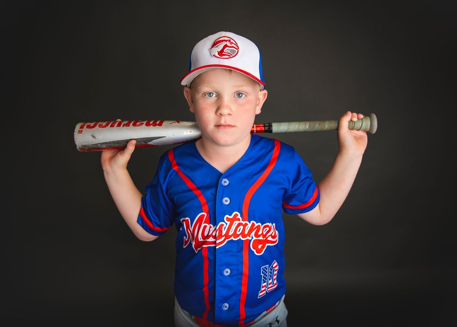 Young boy in blue Mustangs baseball jersey holding bat across shoulders in studio portrait.