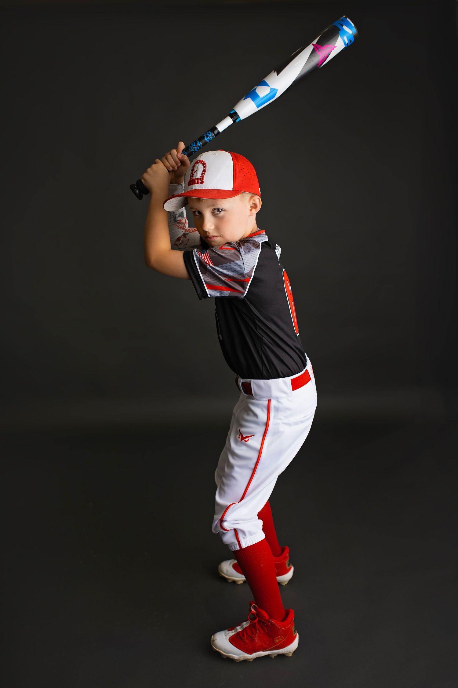 Young baseball player in red Colts uniform swinging bat in batting stance on dark background.