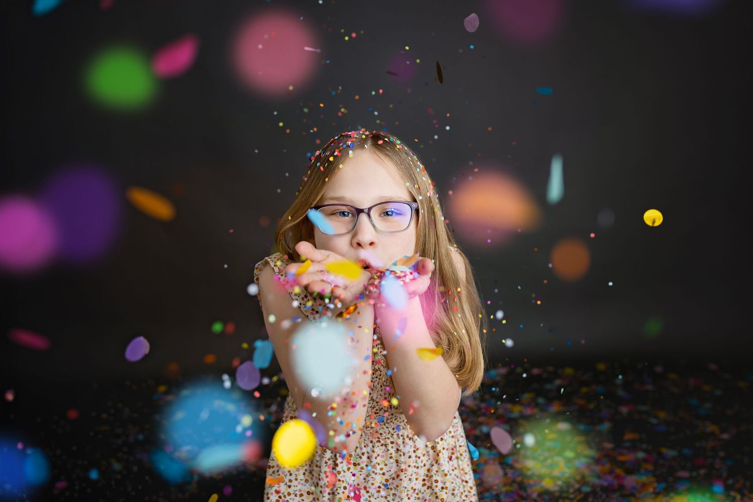 Young girl with glasses blowing colorful confetti from her hands in a festive studio portrait.