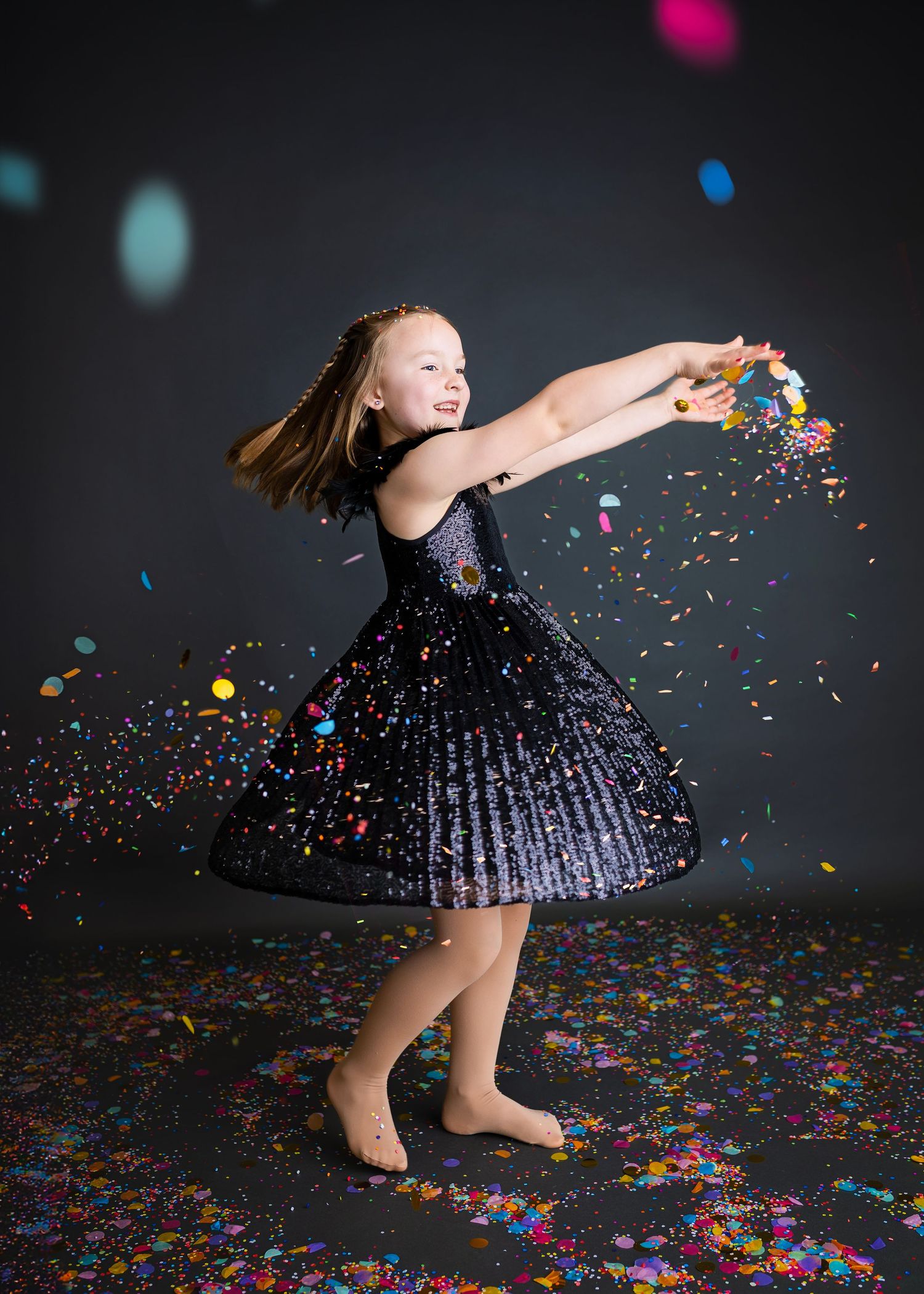 Joyful girl in black sparkle dress spinning while tossing colorful confetti around.