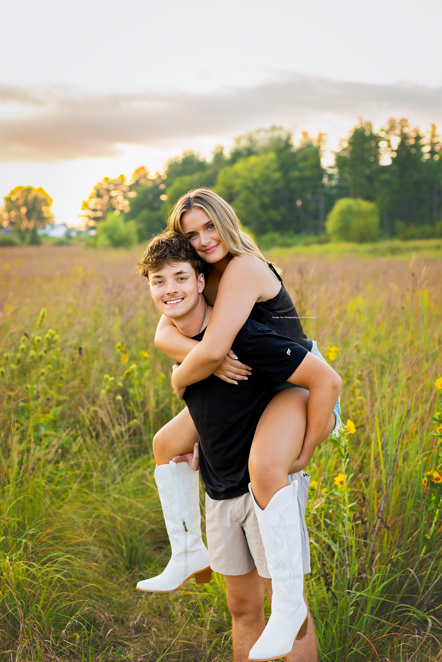 A young couple poses in a sunny meadow, the boy giving the girl a piggyback ride as she wears white cowboy boots.