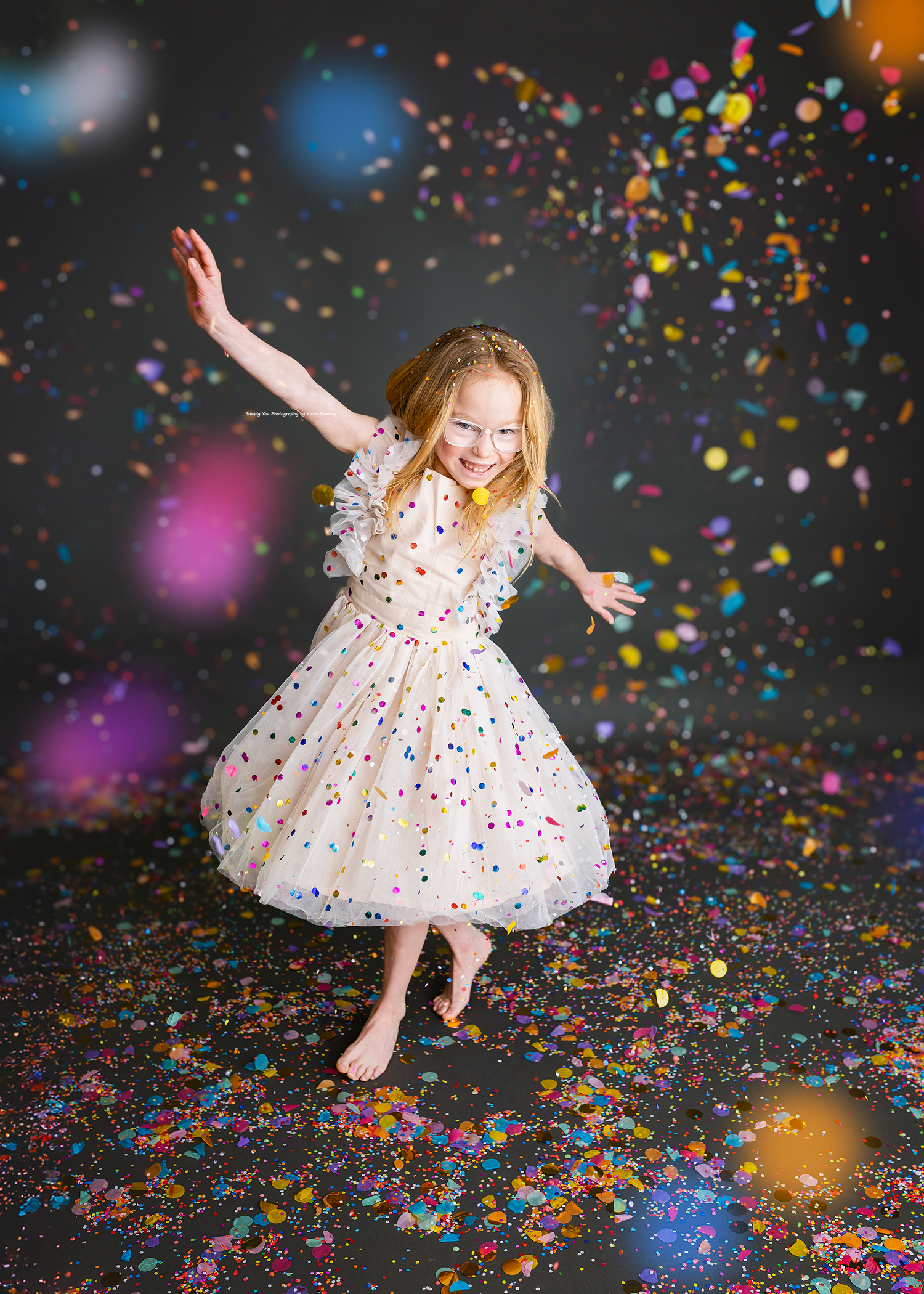 Joyful young girl in white polka dot dress twirls barefoot as vibrant confetti rains down around her.
