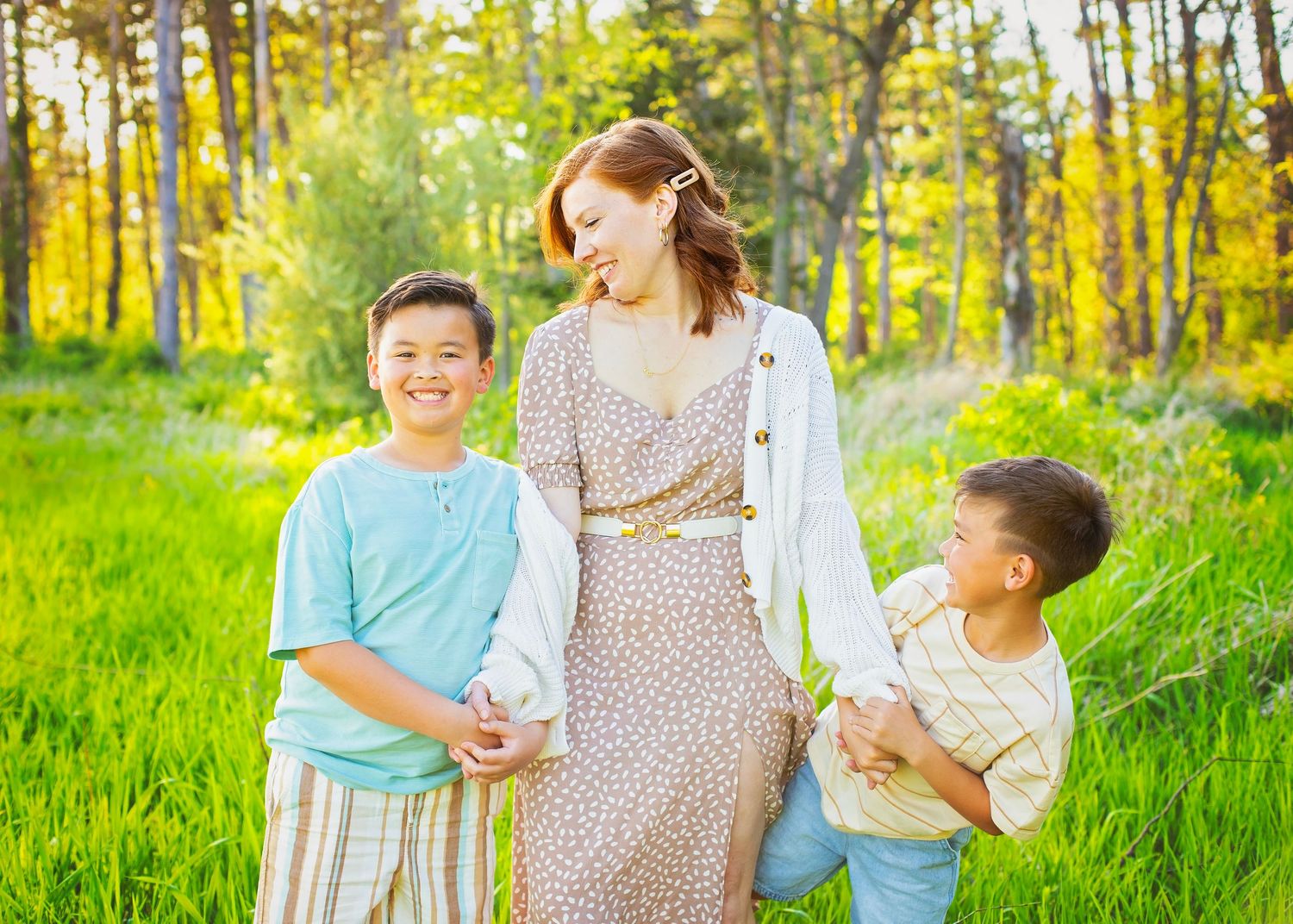 A woman in a floral dress walks hand-in-hand with two boys through a sunny green meadow.