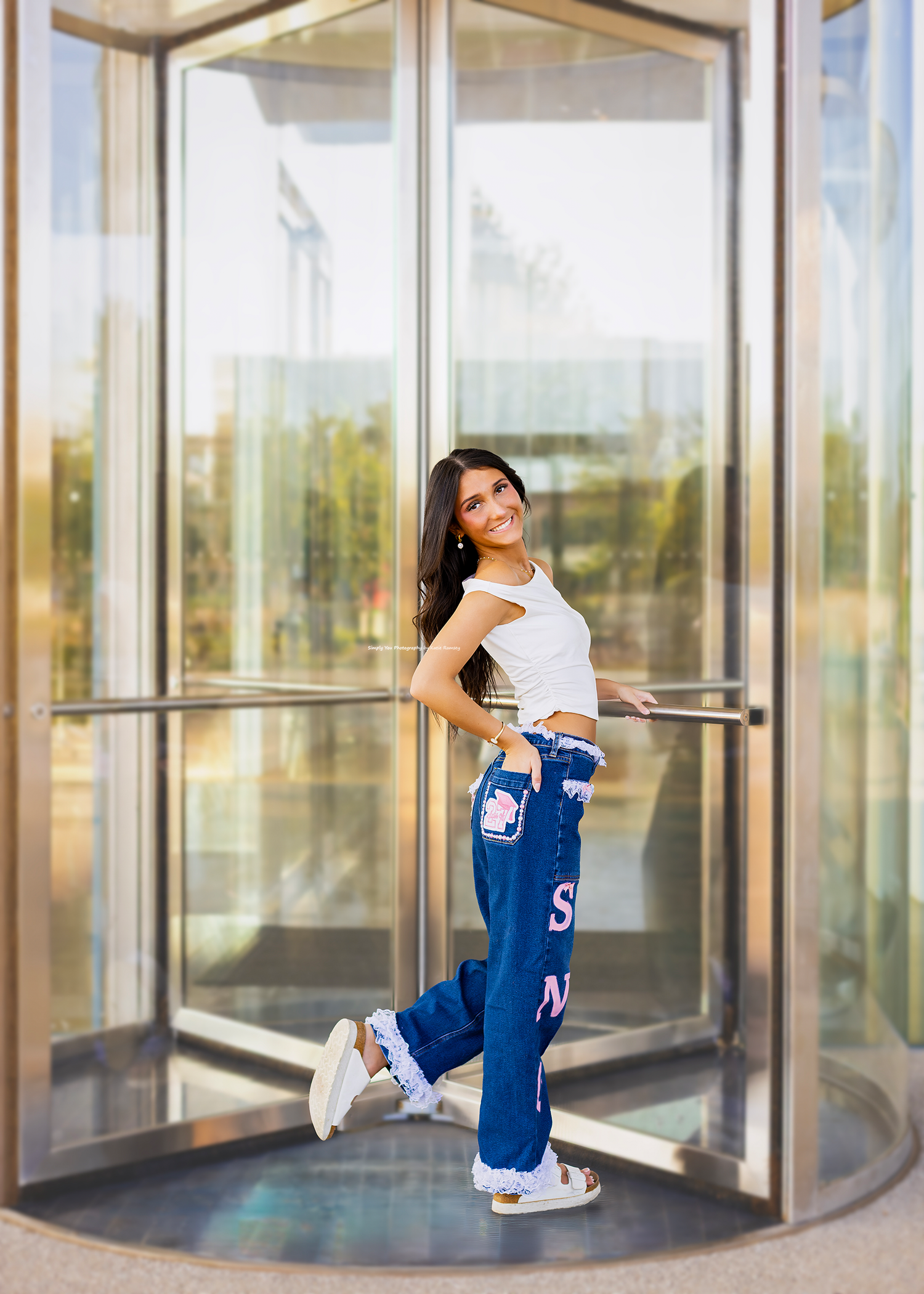 Young woman in white crop top and blue graphic jeans posing at a revolving glass door entrance.