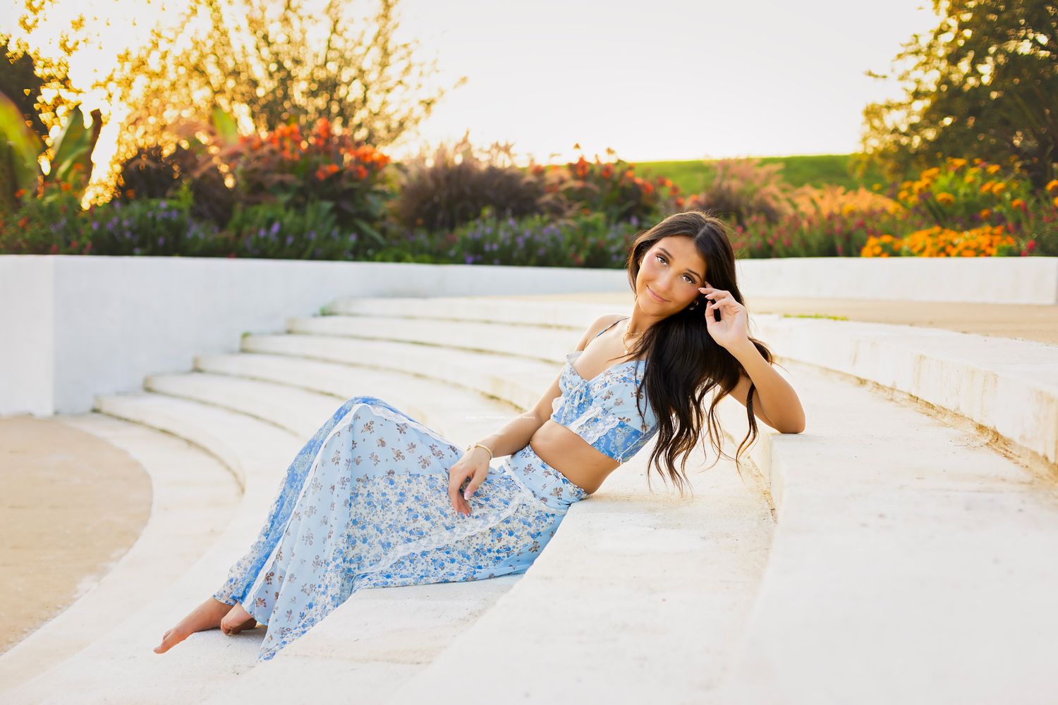 Young woman in blue floral two-piece outfit sitting relaxed on white stone steps in garden setting.