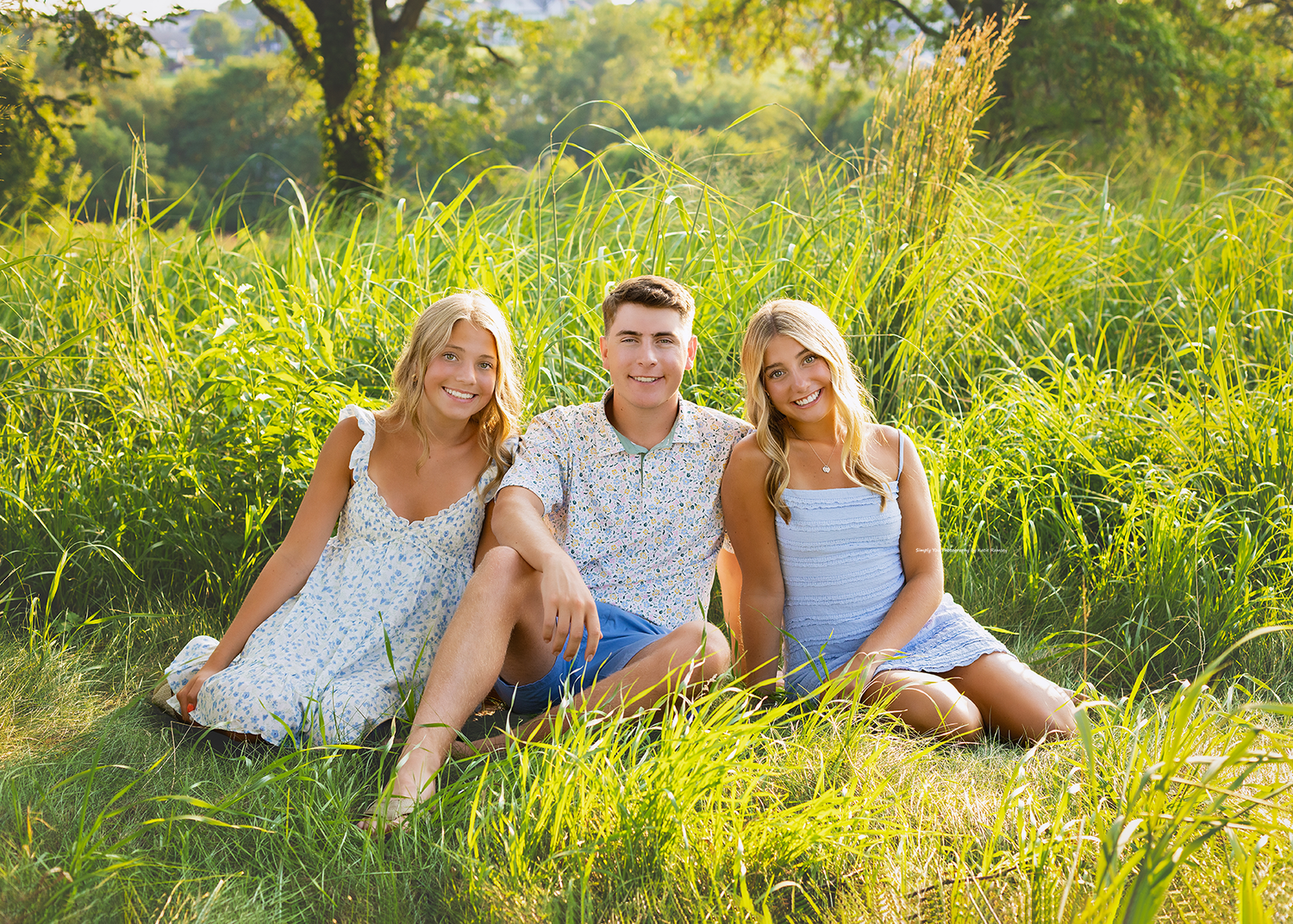 Three smiling teens sitting in lush green grass outdoors on a sunny day, two blonde girls flanking a young man.