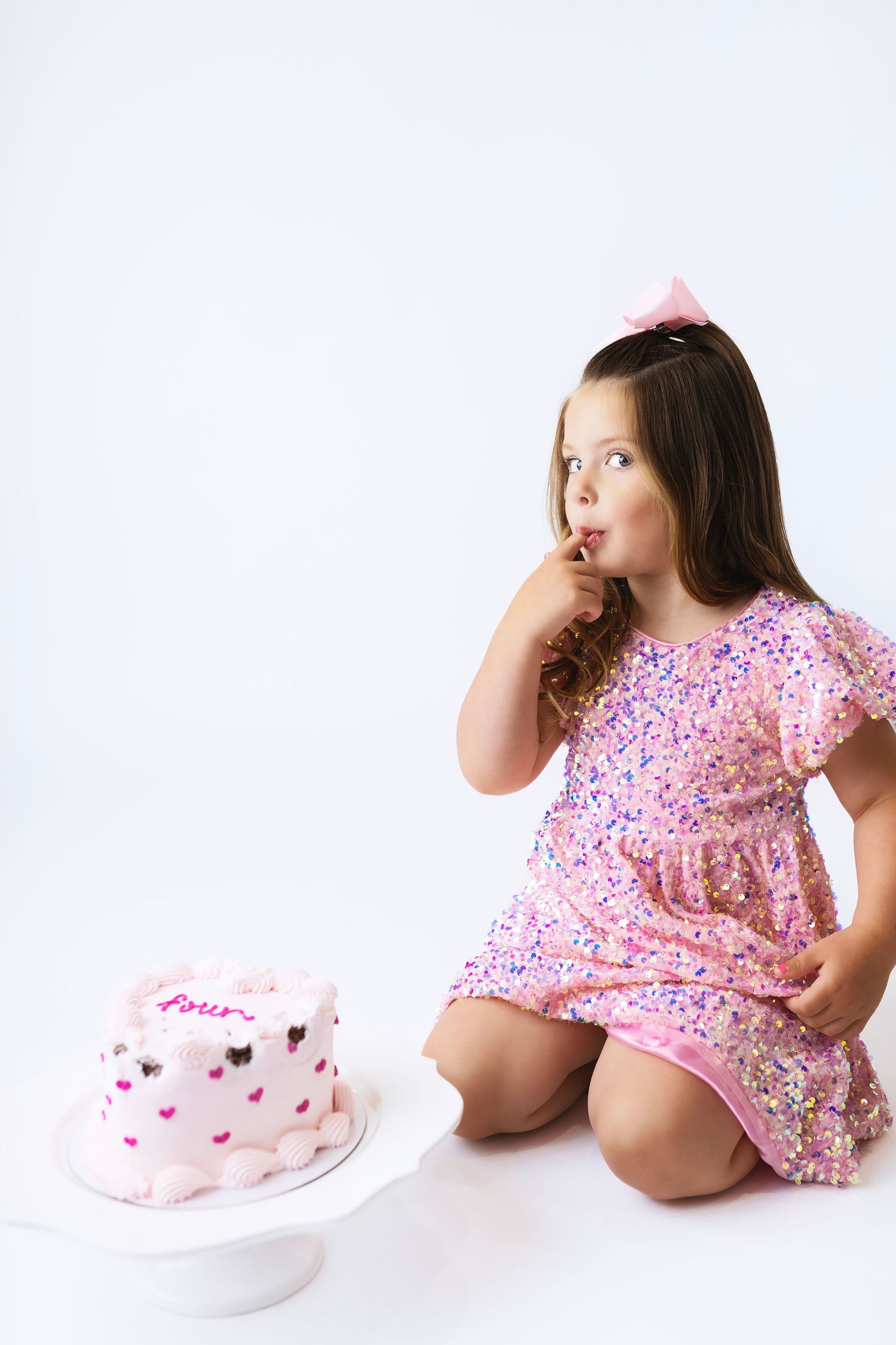 Young girl in pink sequin dress licks finger while kneeling beside a pink birthday cake on a white cake stand.