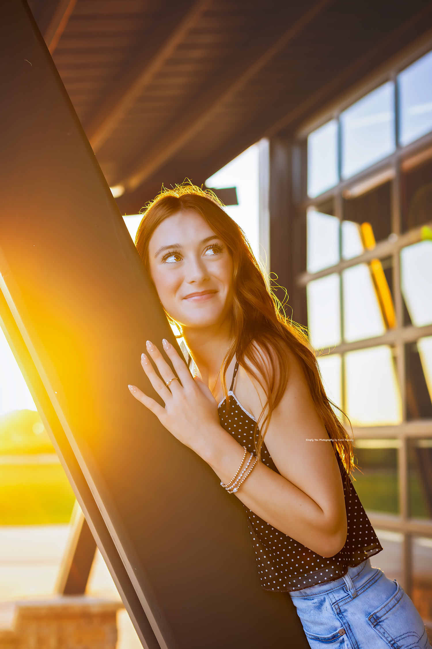 Young woman in polka dot top and jeans leans against a dark pillar, bathed in warm golden sunset light.