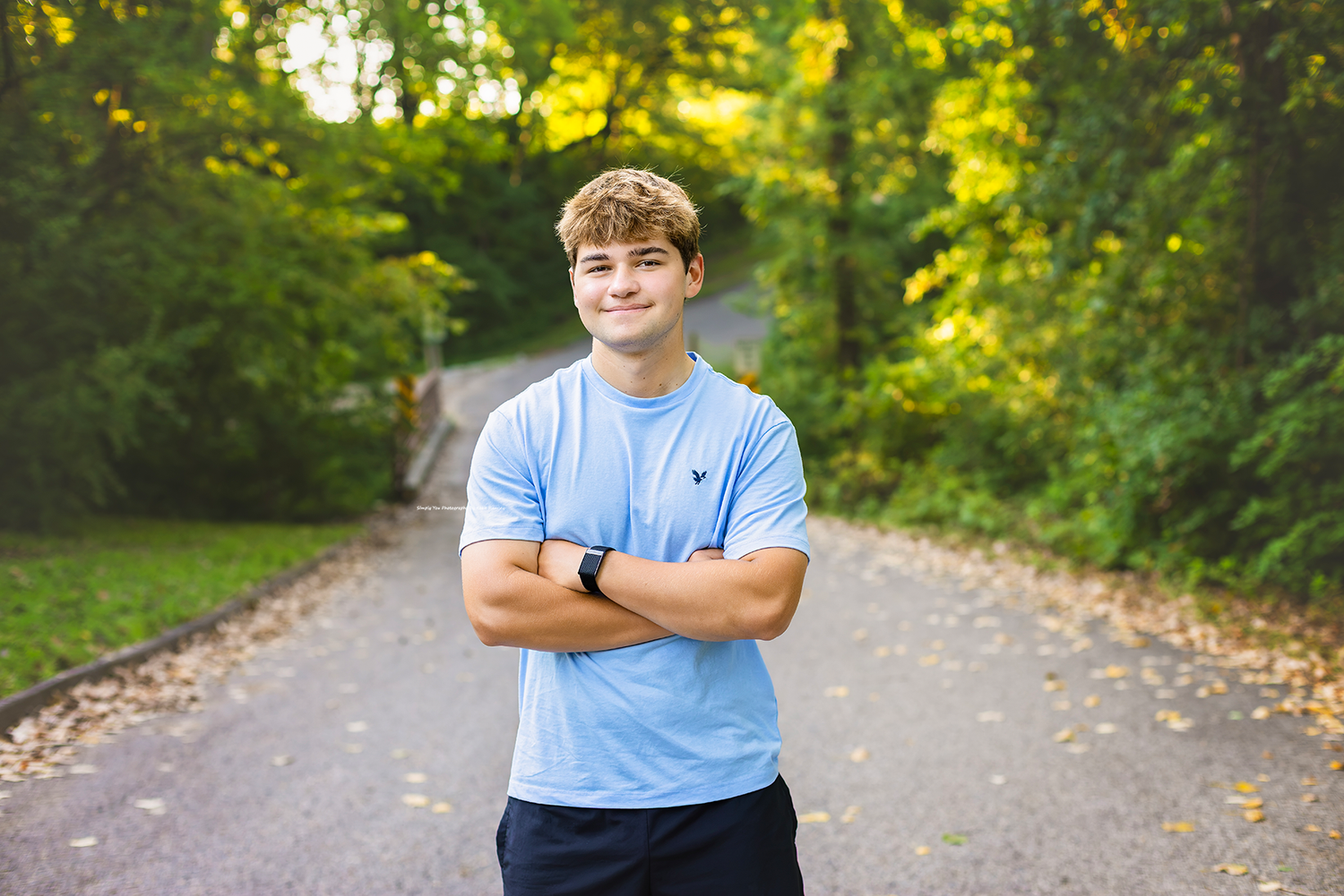 A smiling young man in a light blue shirt stands with arms crossed on a tree-lined path in autumn.