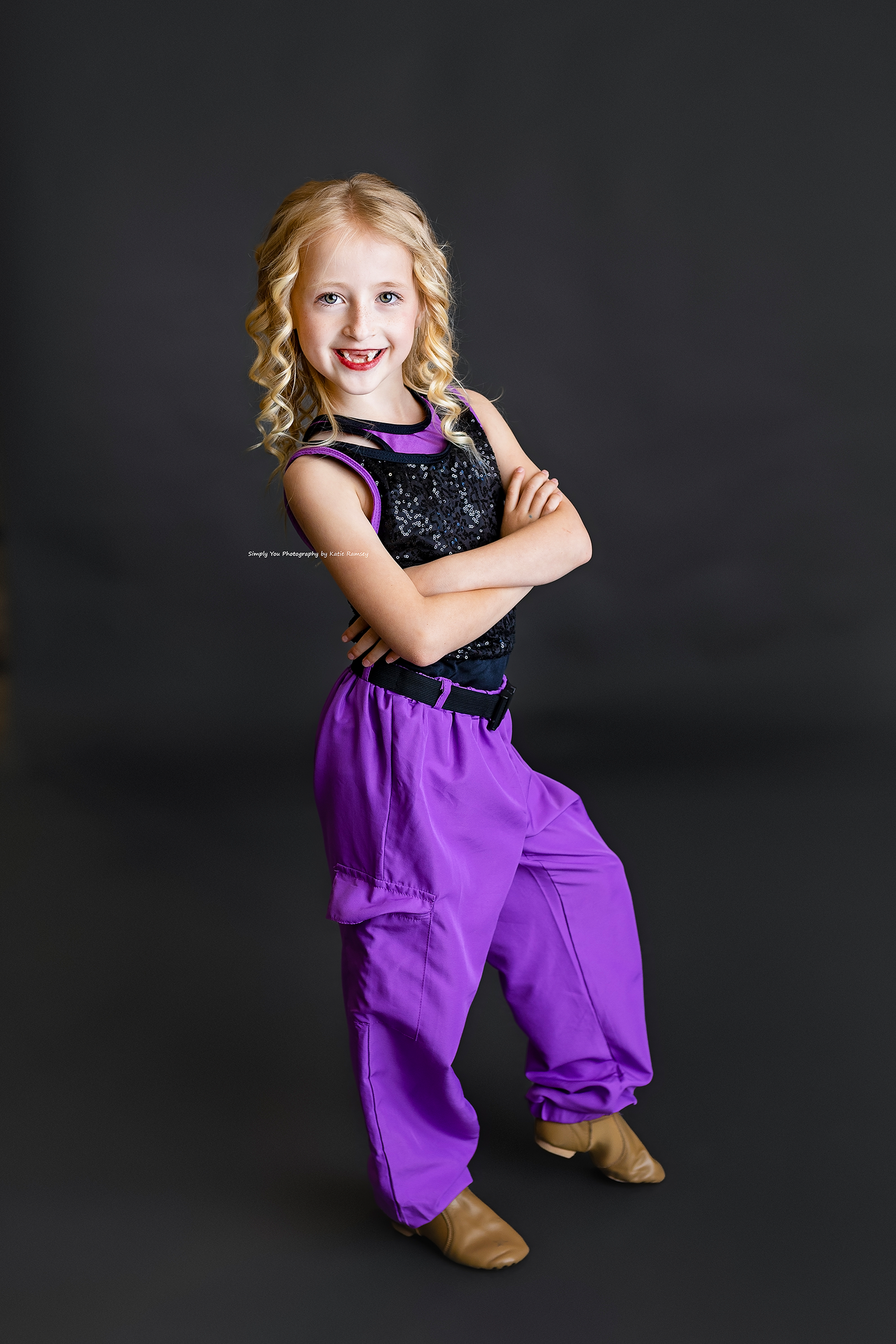 Young blonde girl in purple harem pants and black sequin top poses confidently with arms crossed in dance portrait.