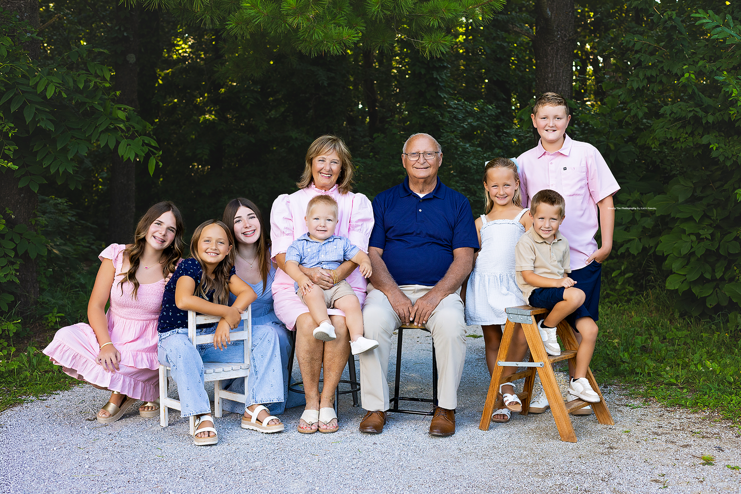 Large family portrait with grandparents and grandchildren smiling outdoors near lush green trees.