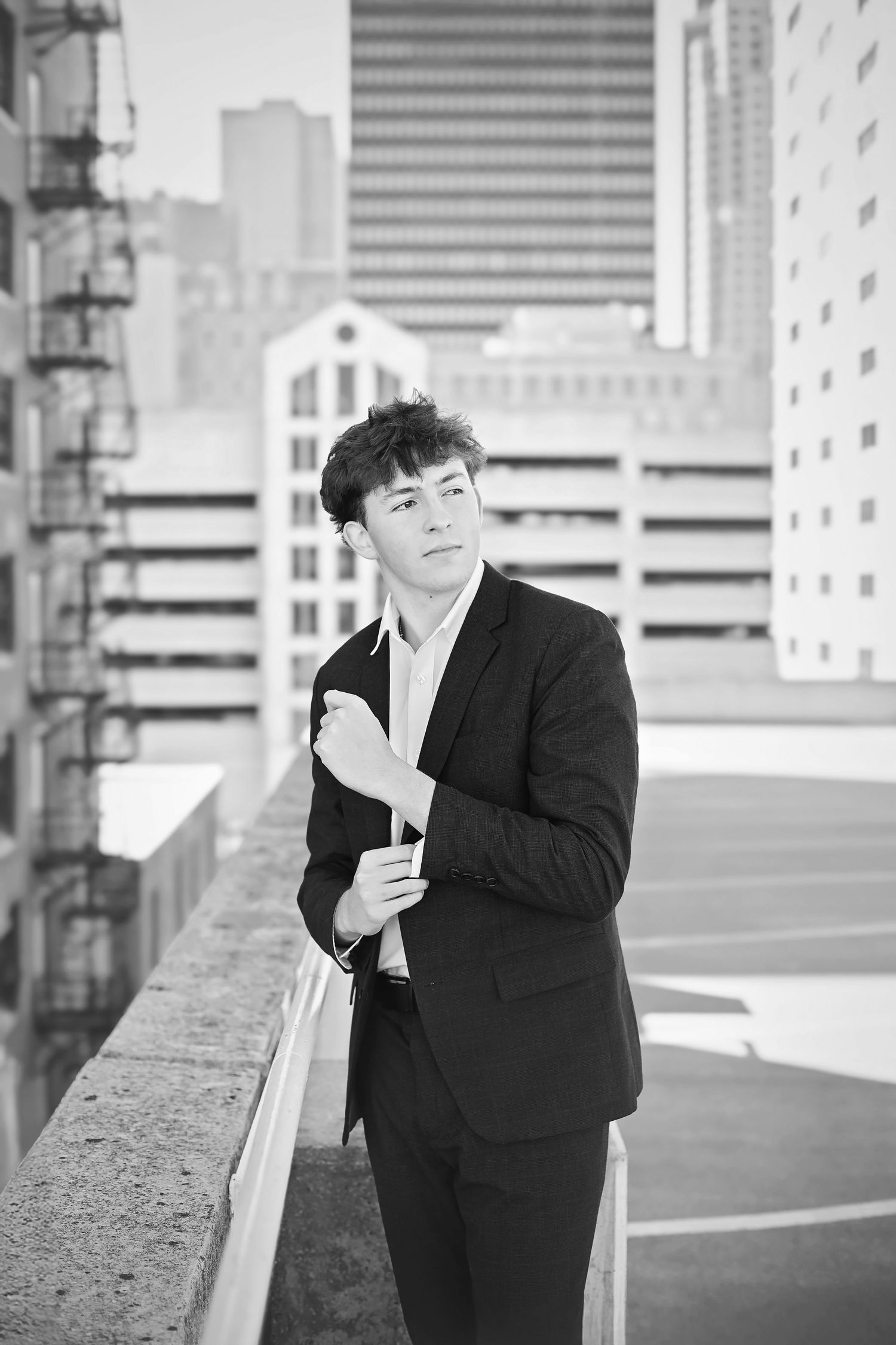 Young man in black suit poses on urban rooftop with city skyline, black and white.