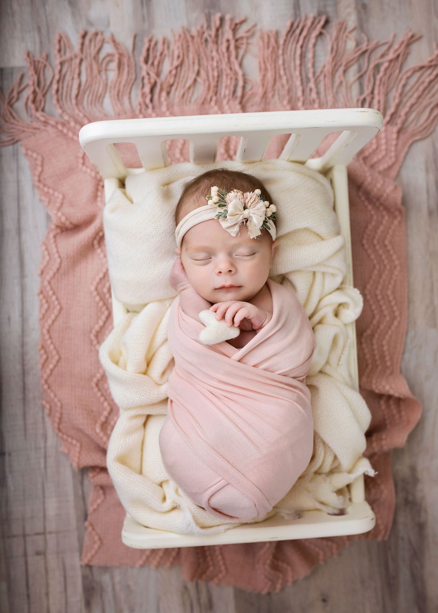 Sleeping newborn baby girl in pink swaddle with bow headband in white miniature crib on pink blanket.