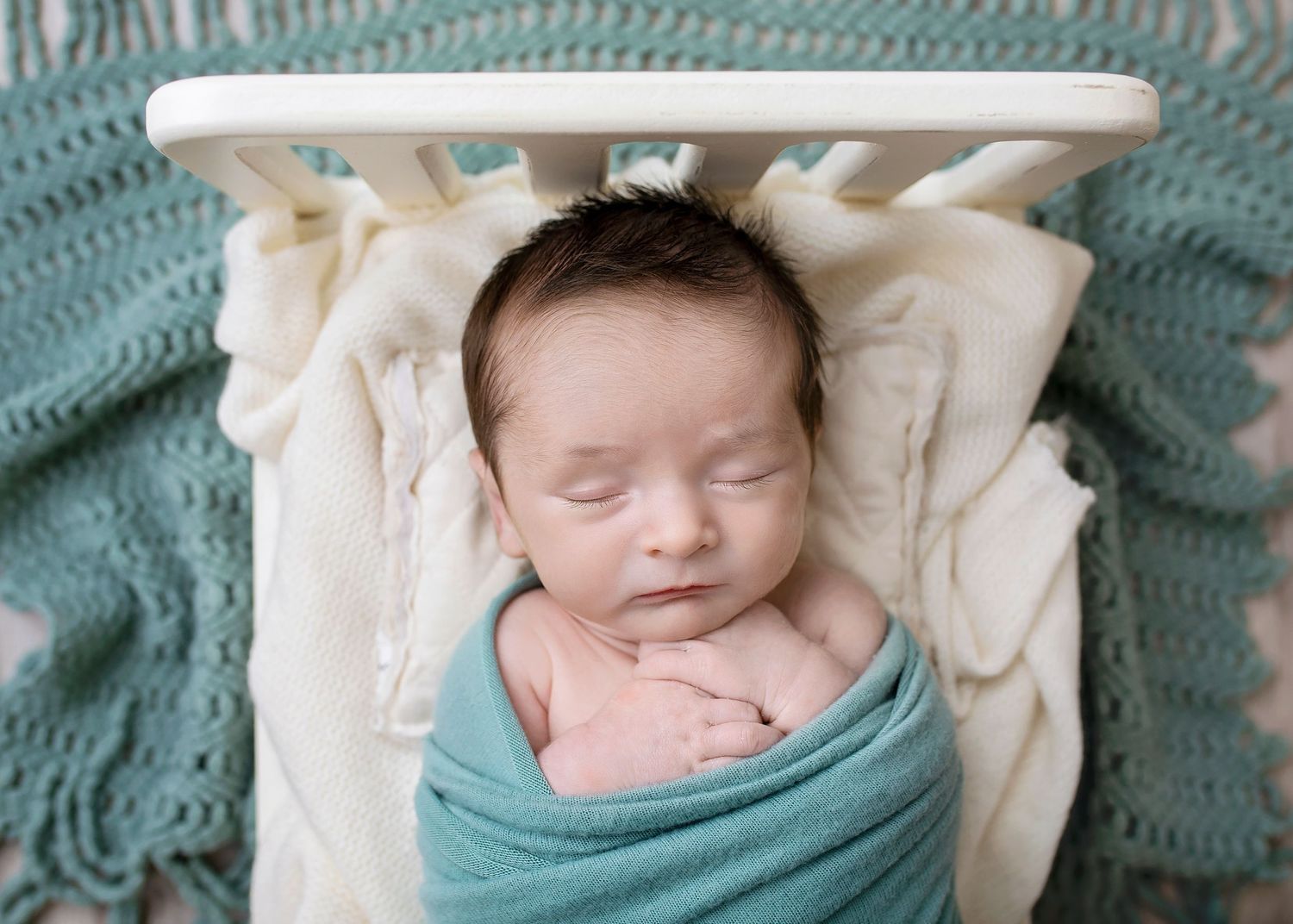 Sleeping newborn baby swaddled in teal wrap resting in a small white wooden bed with cream blanket.