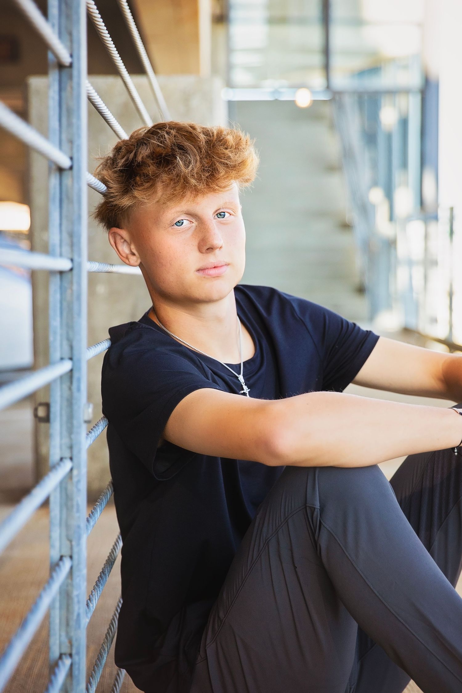 Teen boy in black t-shirt sitting against metal railing on concrete steps looking at camera.