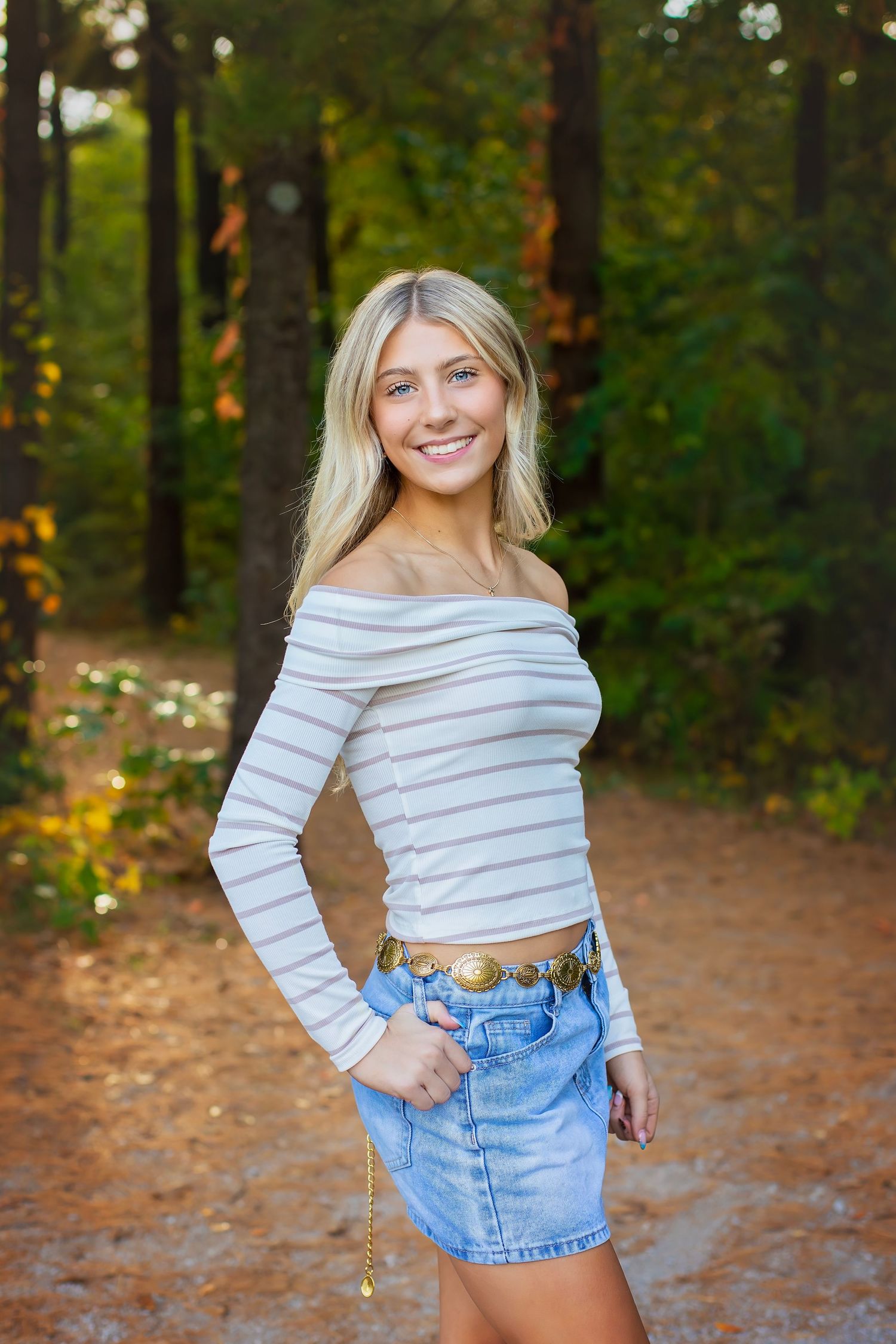 A smiling blonde woman poses in a striped off-shoulder top and denim skirt on a forest path.