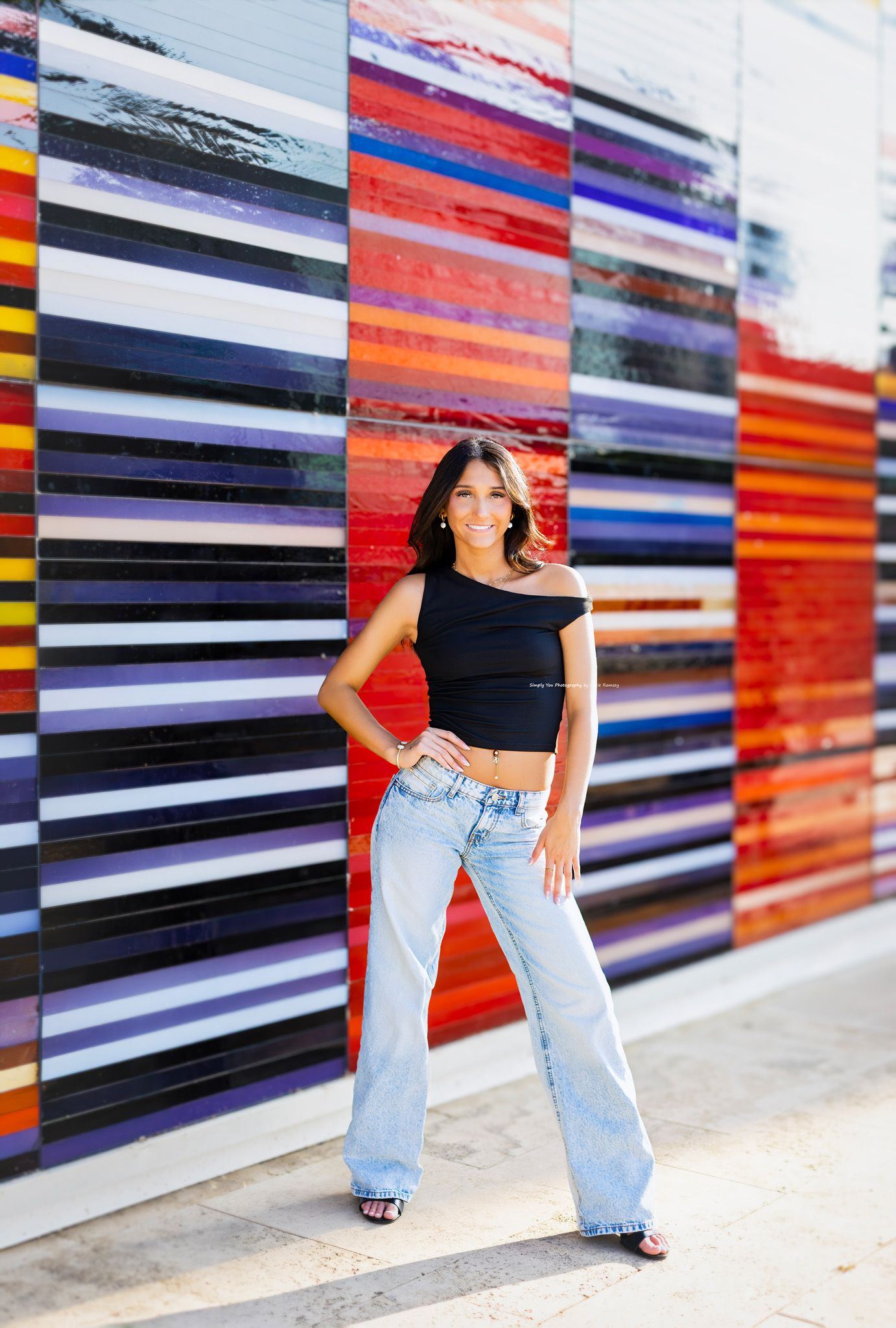 Young woman in black crop top and wide-leg jeans posing confidently in front of colorful striped mural wall.