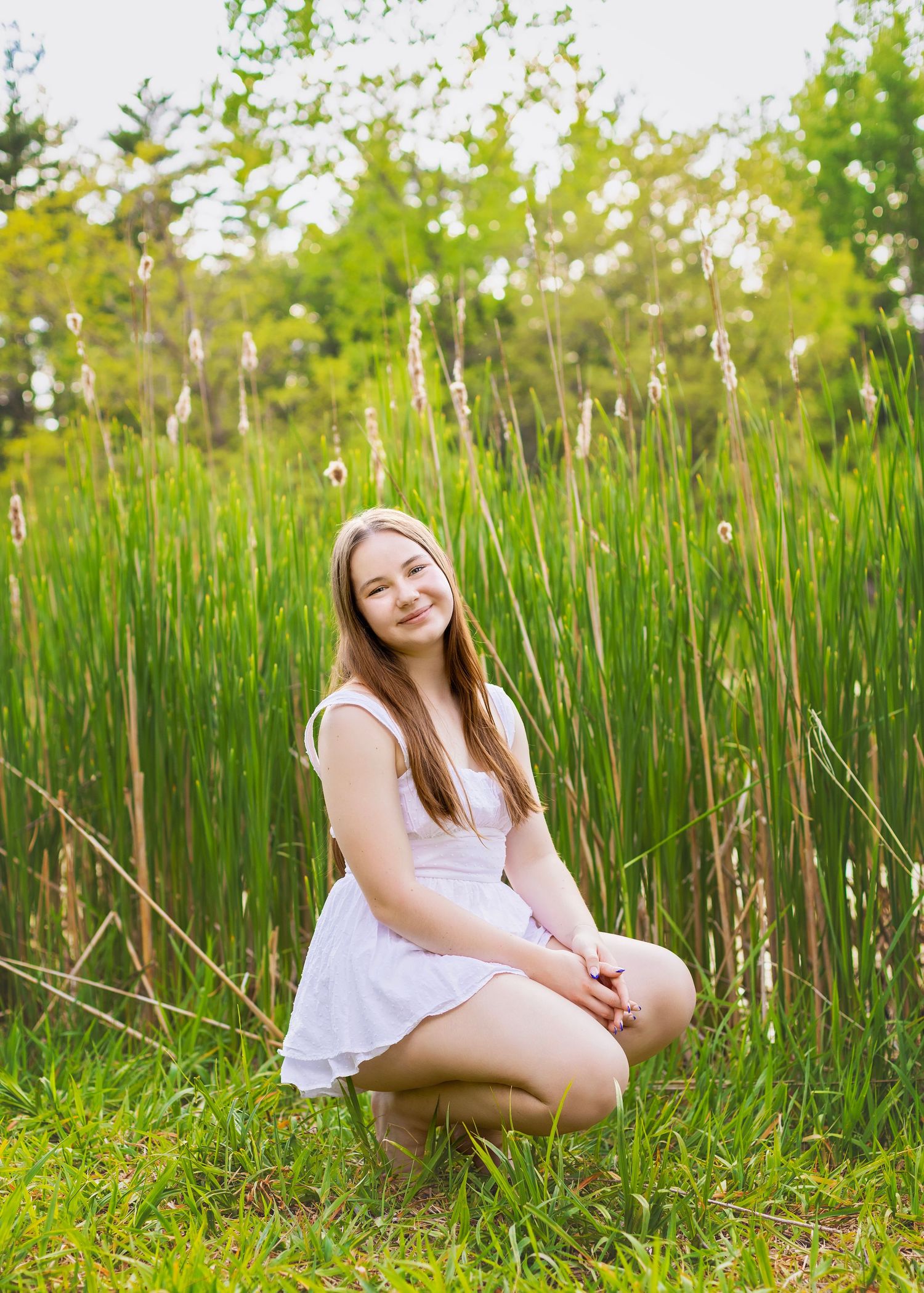 Smiling teenage girl in white dress crouching in tall green grass in a sunny outdoor setting.
