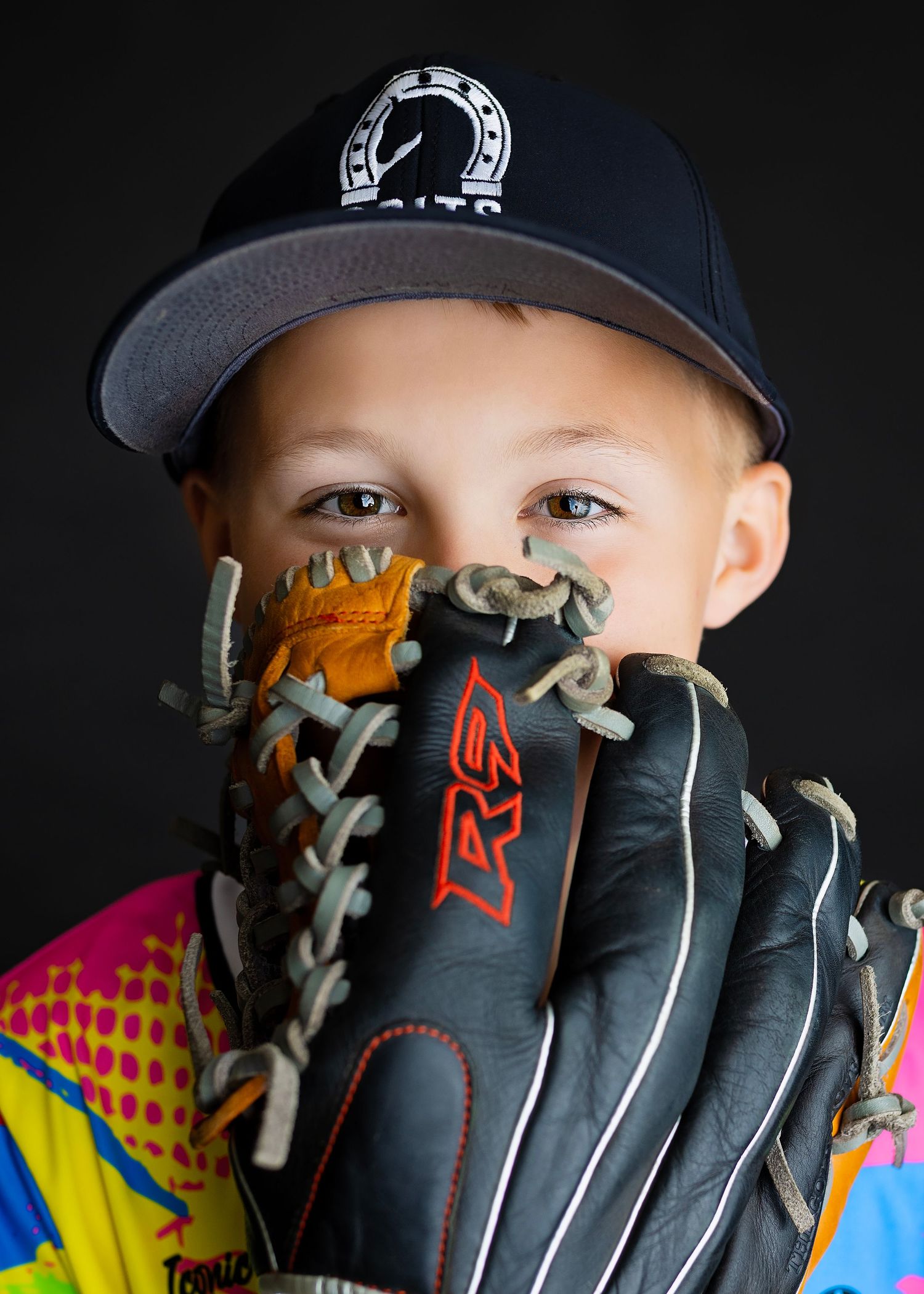 Young baseball player in colorful jersey holds leather RAW glove up to face wearing black cap against dark background.