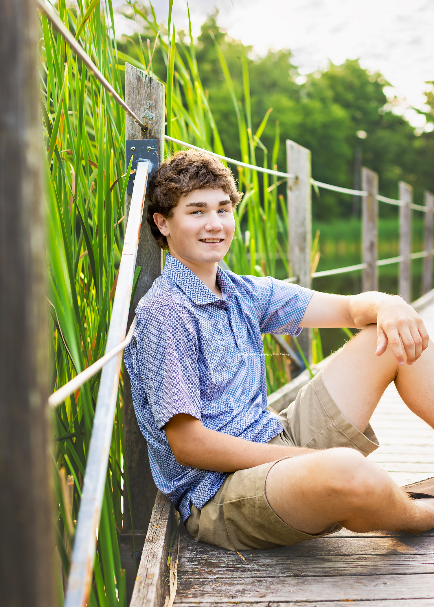 A smiling teenage boy sits relaxed on a wooden dock surrounded by tall green reeds on a sunny day.