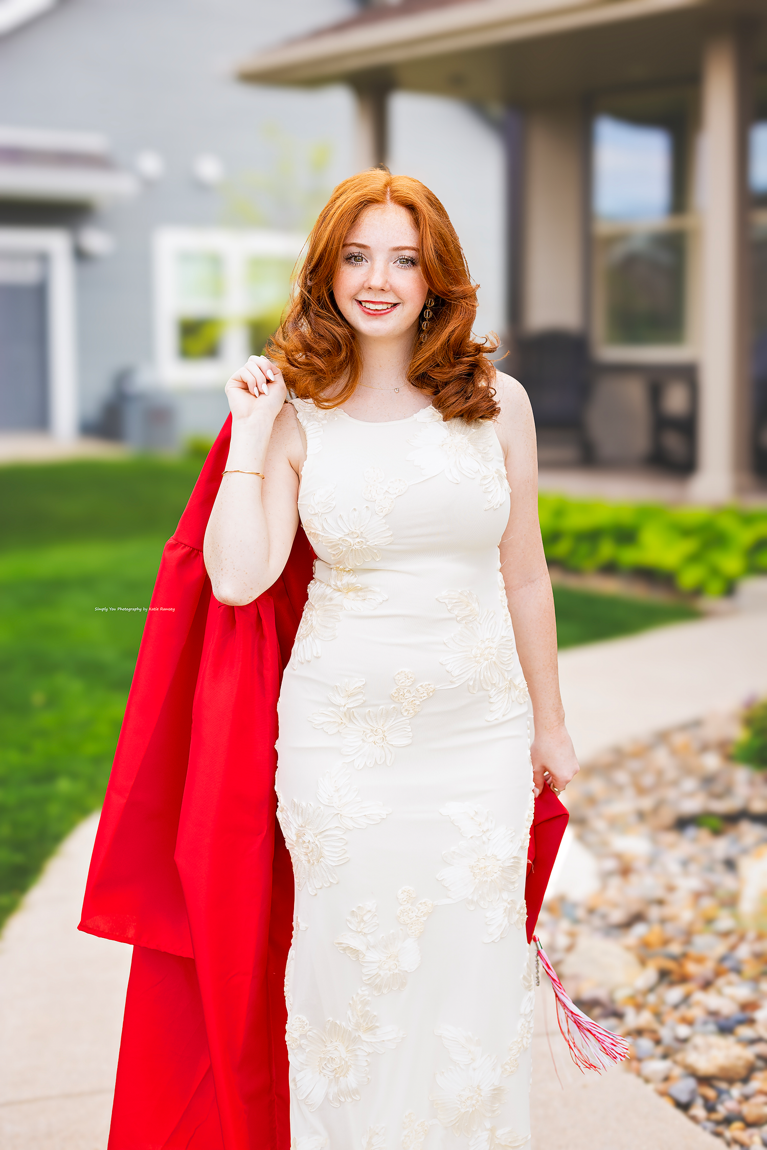 Red-haired woman in white lace gown holding red graduation gown outside a suburban home.
