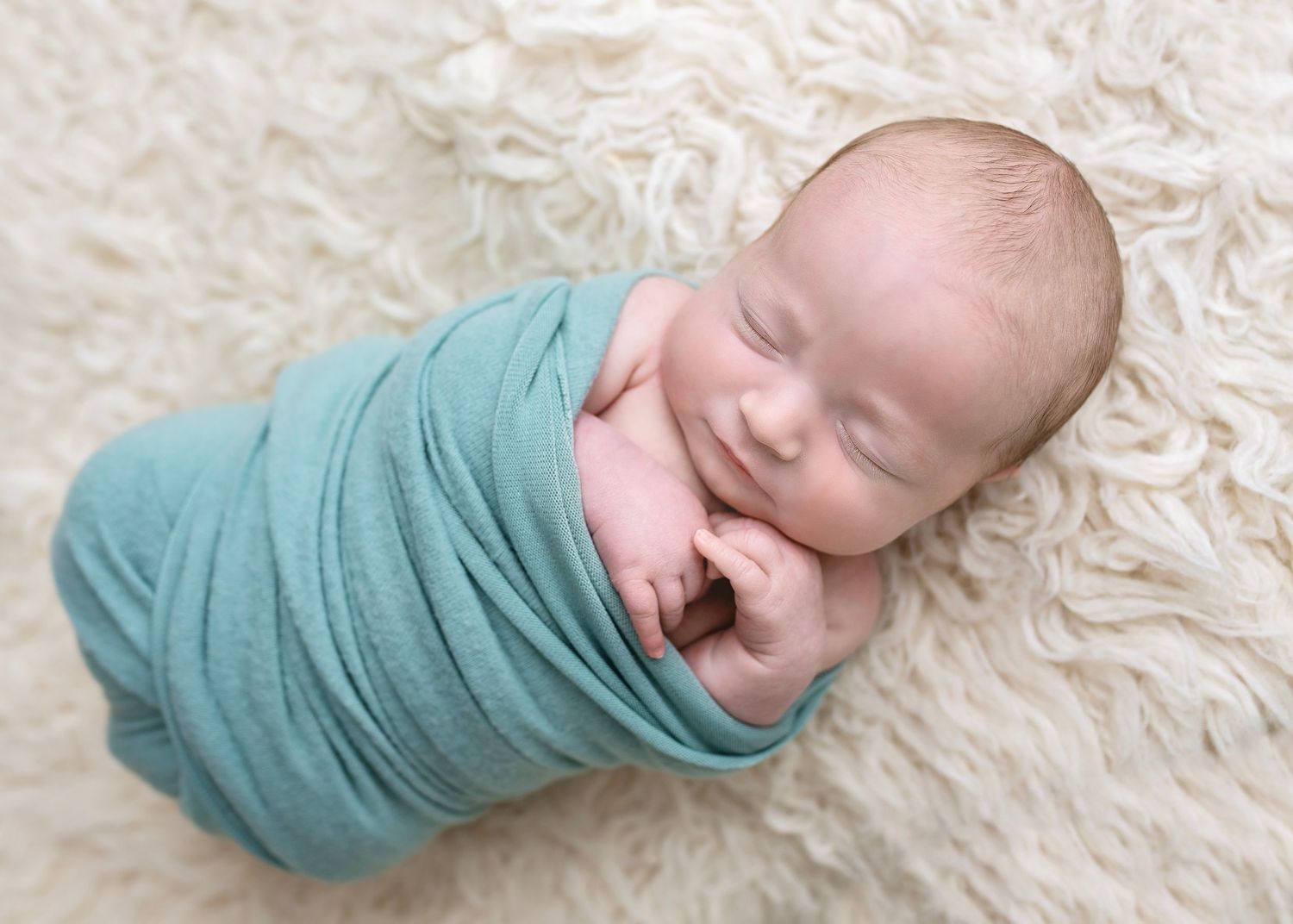 Smiling sleeping newborn baby tightly swaddled in teal wrap on fluffy cream fur blanket.