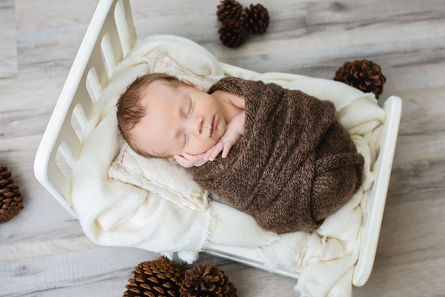 Smiling newborn baby wrapped in brown knit swaddle sleeping in small white wooden crib with pinecones.