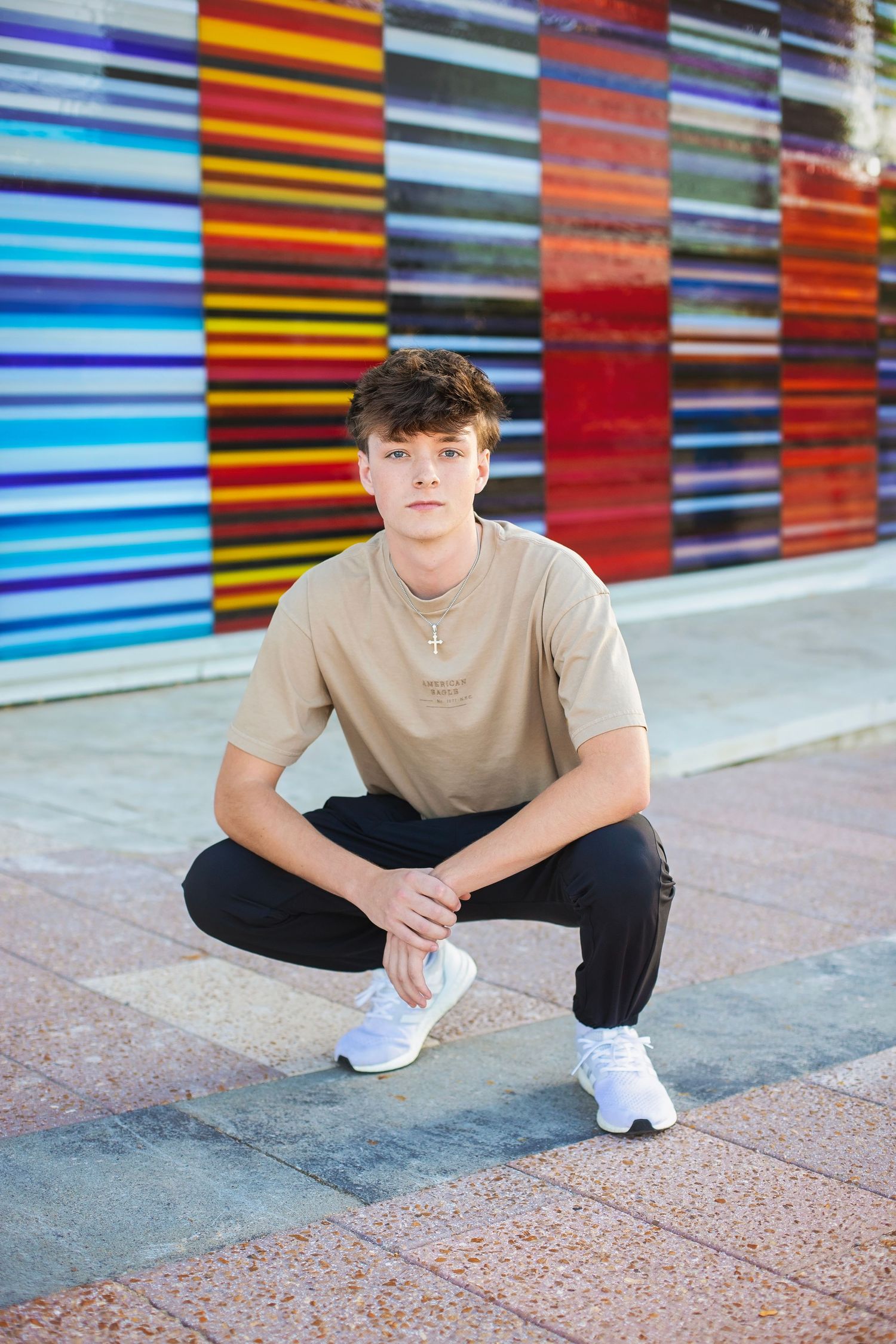 Teen boy in beige t-shirt crouching in front of colorful striped wall outdoors.
