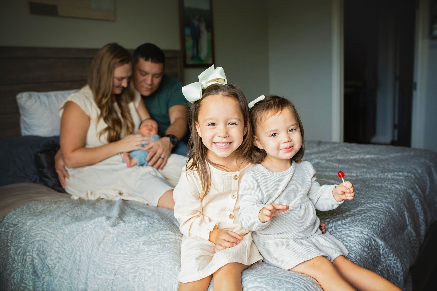 Two young sisters smiling on a bed while parents play with a newborn in the background.