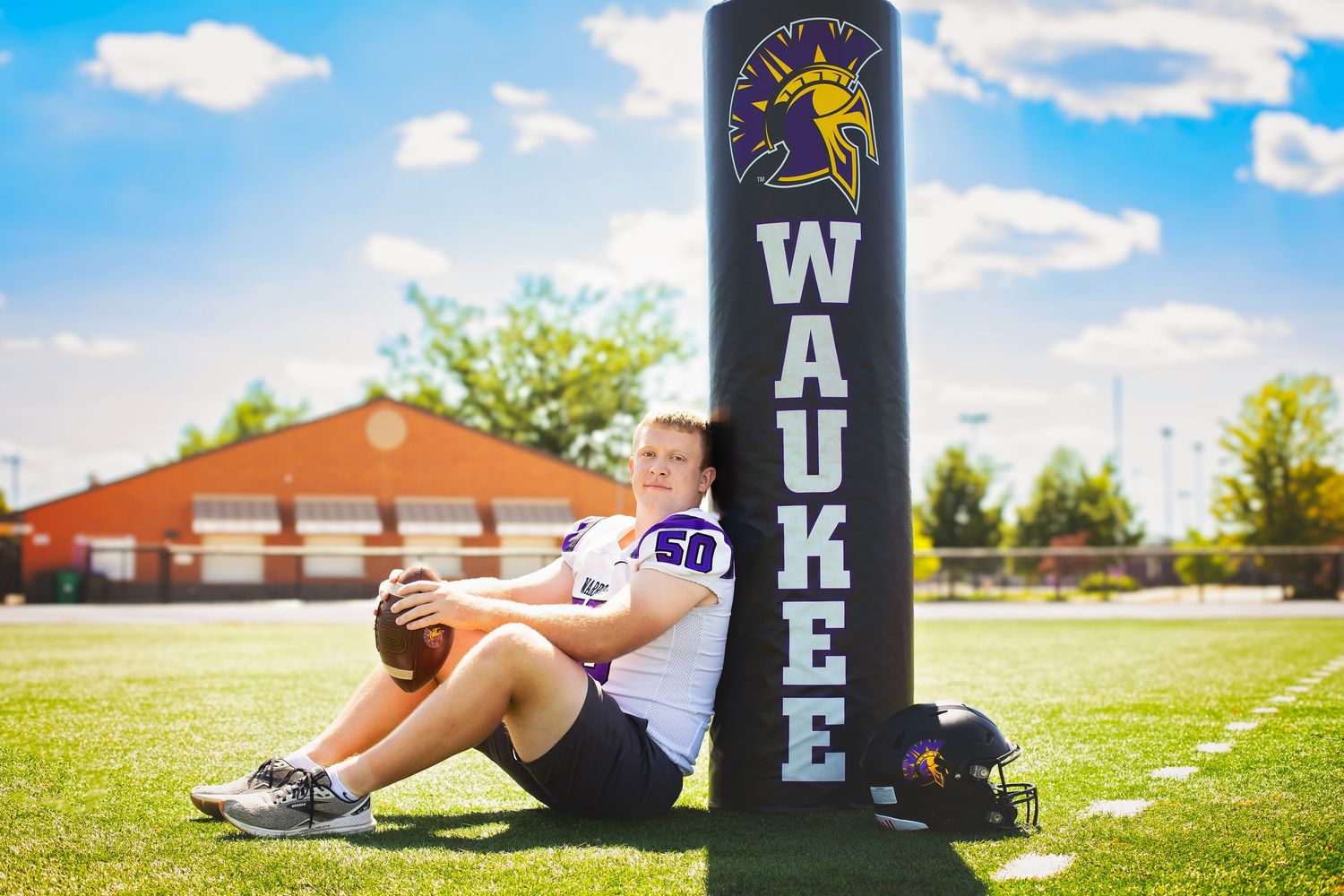 Football player sits against Waukee branded field marker holding a football outdoors.