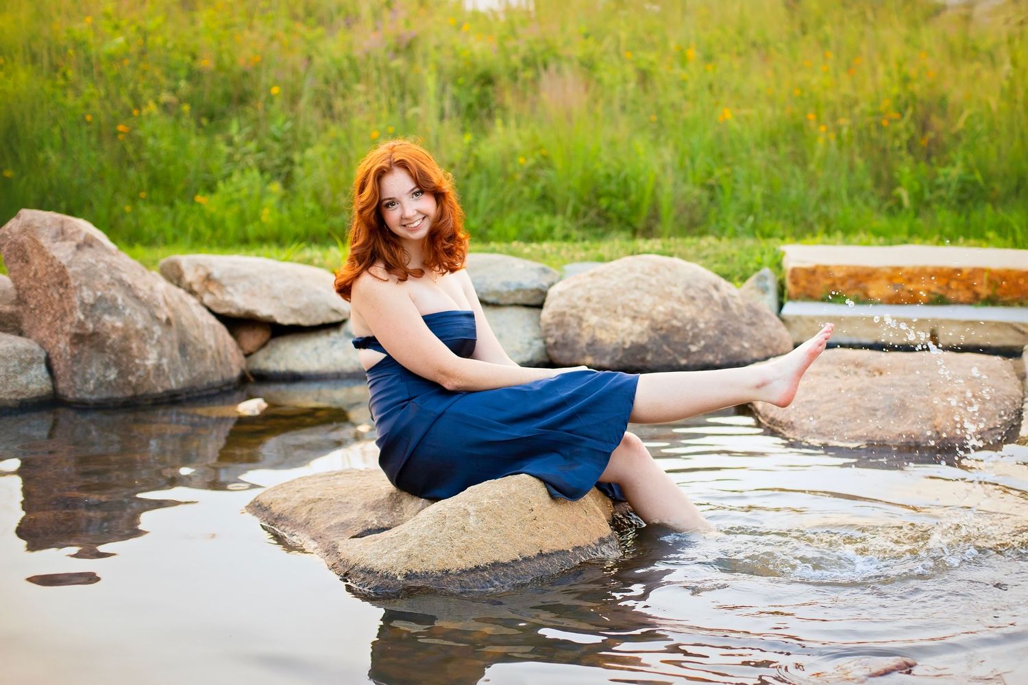 Redhead woman in blue dress sitting on rocks with feet in water surrounded by wildflowers.