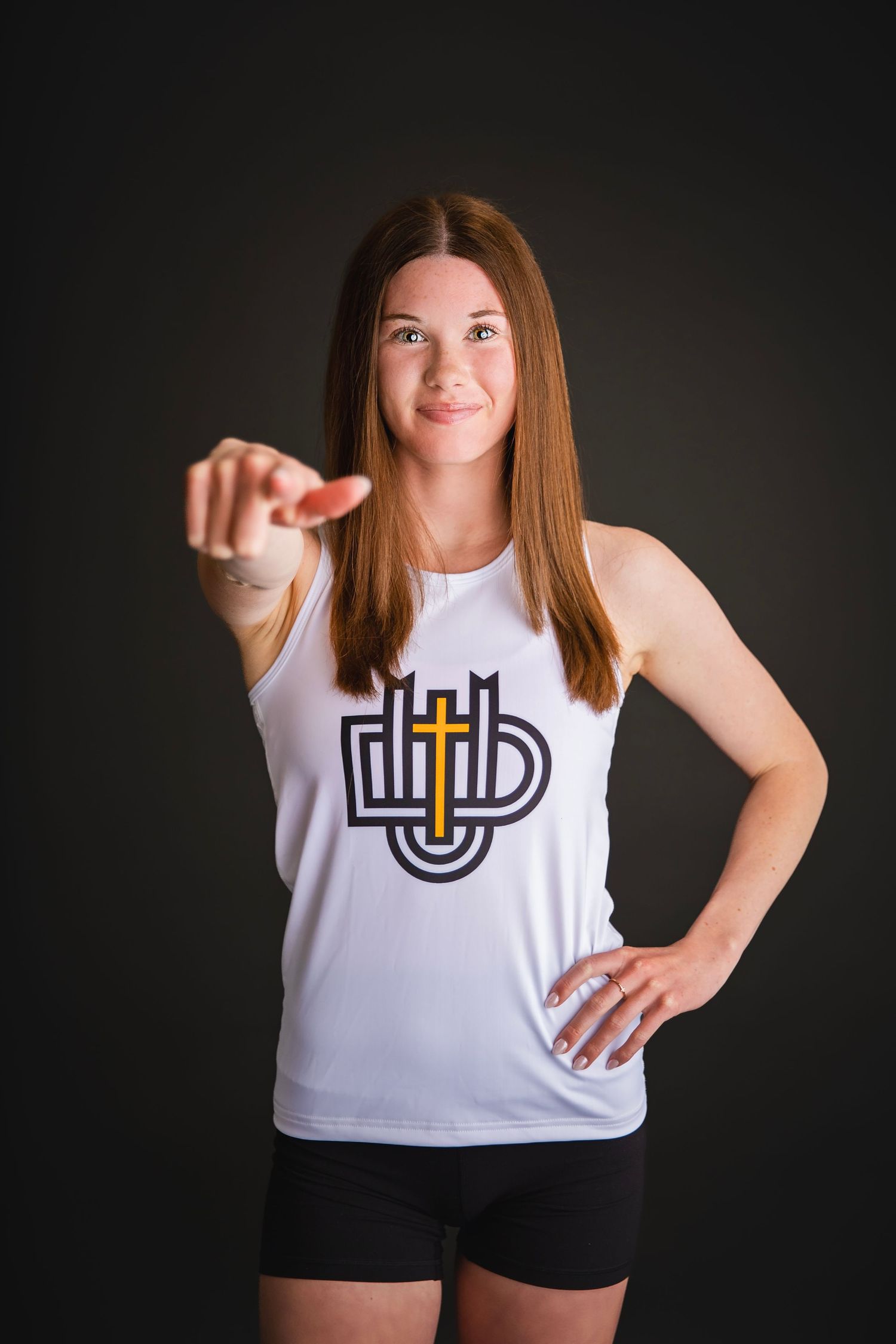 Young female athlete in white tank top with cross logo pointing at camera against dark background.