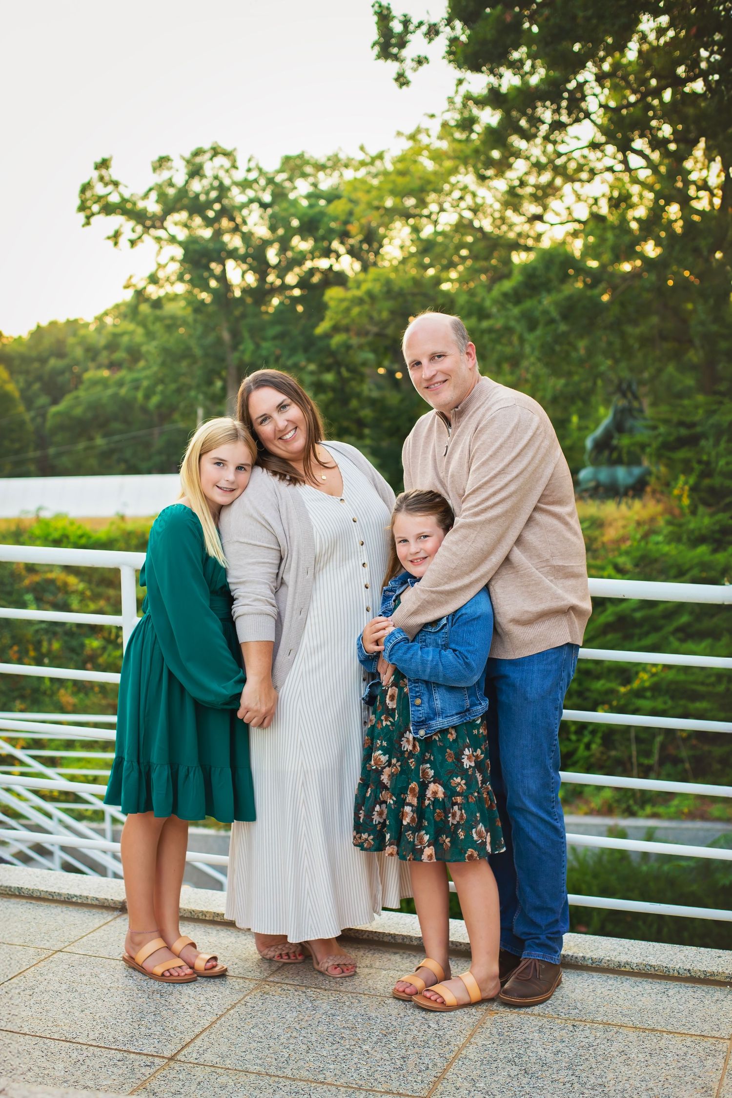 Happy family of four standing together on an outdoor terrace with lush green trees in the background at golden hour.