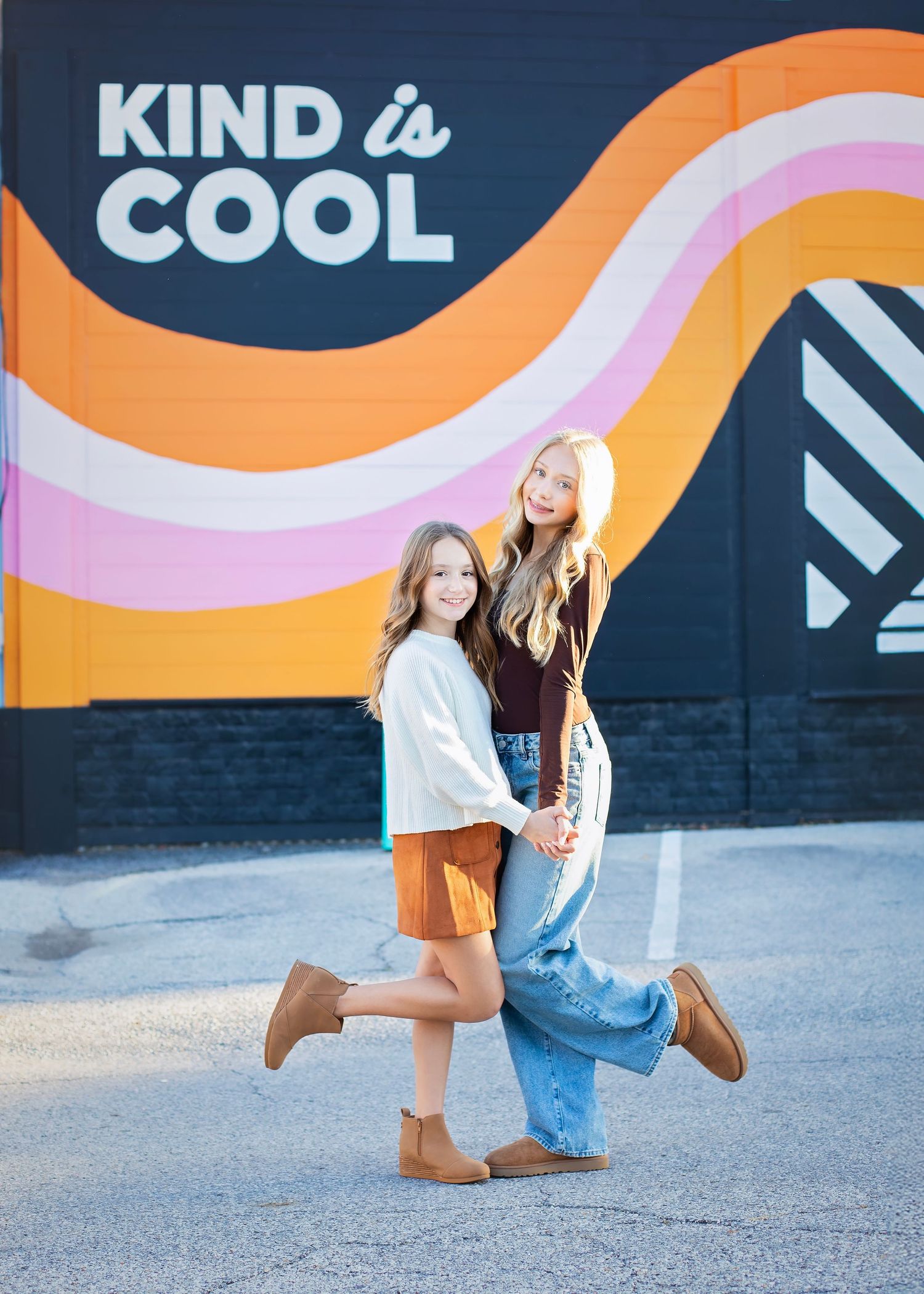 Two girls posing in front of colorful Kind is Cool mural outdoors.