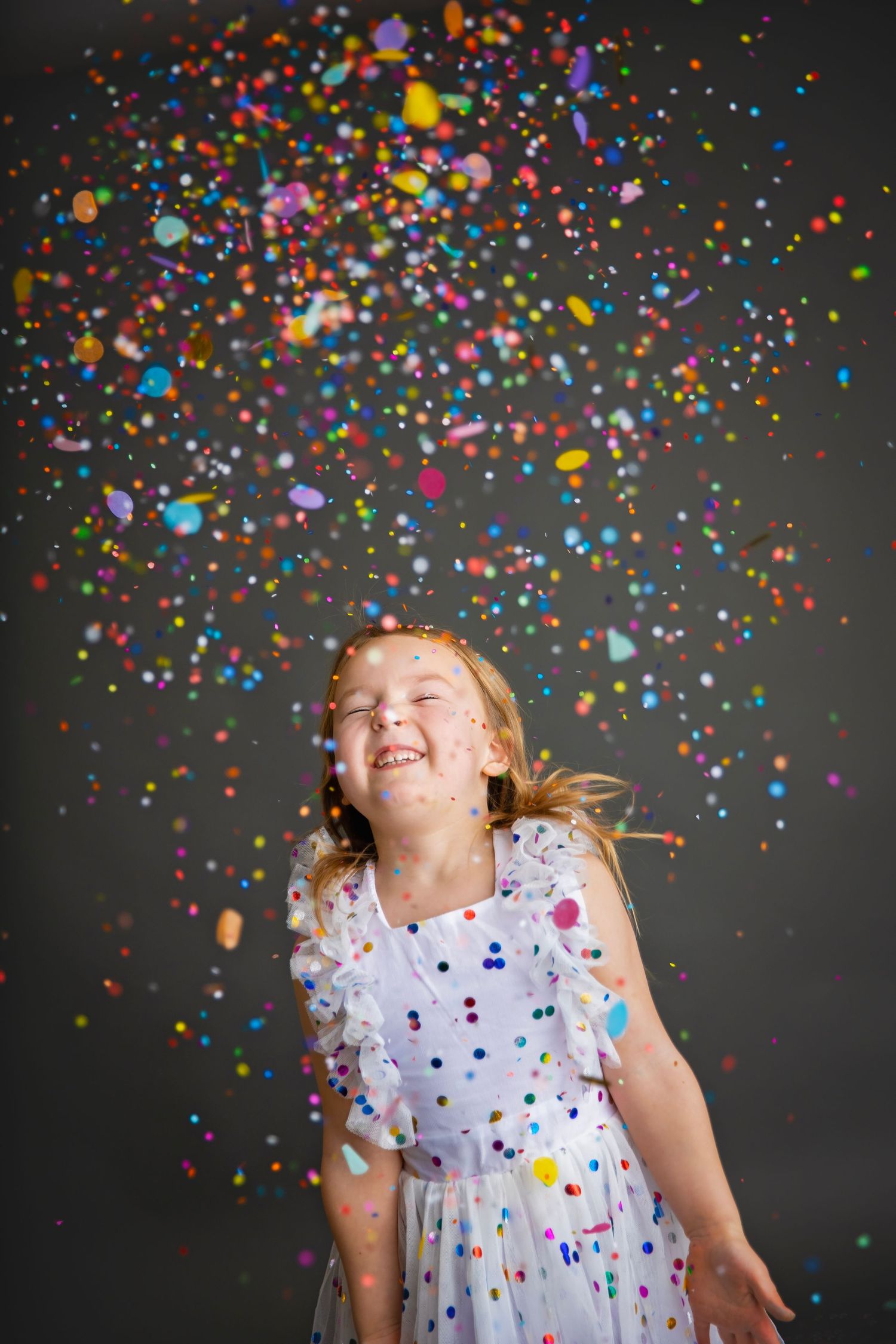 A gleeful young girl in a white dress laughs as colorful confetti rains down around her in a studio setting.