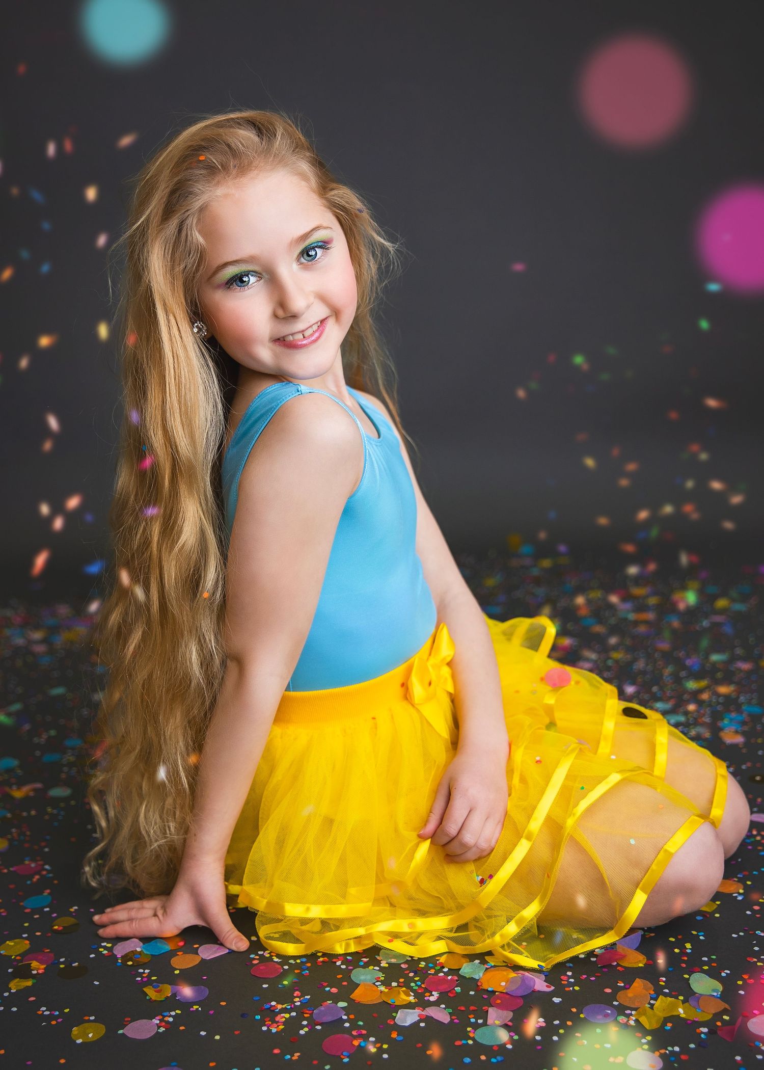 Smiling girl with long hair in blue top and yellow skirt sitting with confetti on gray background.