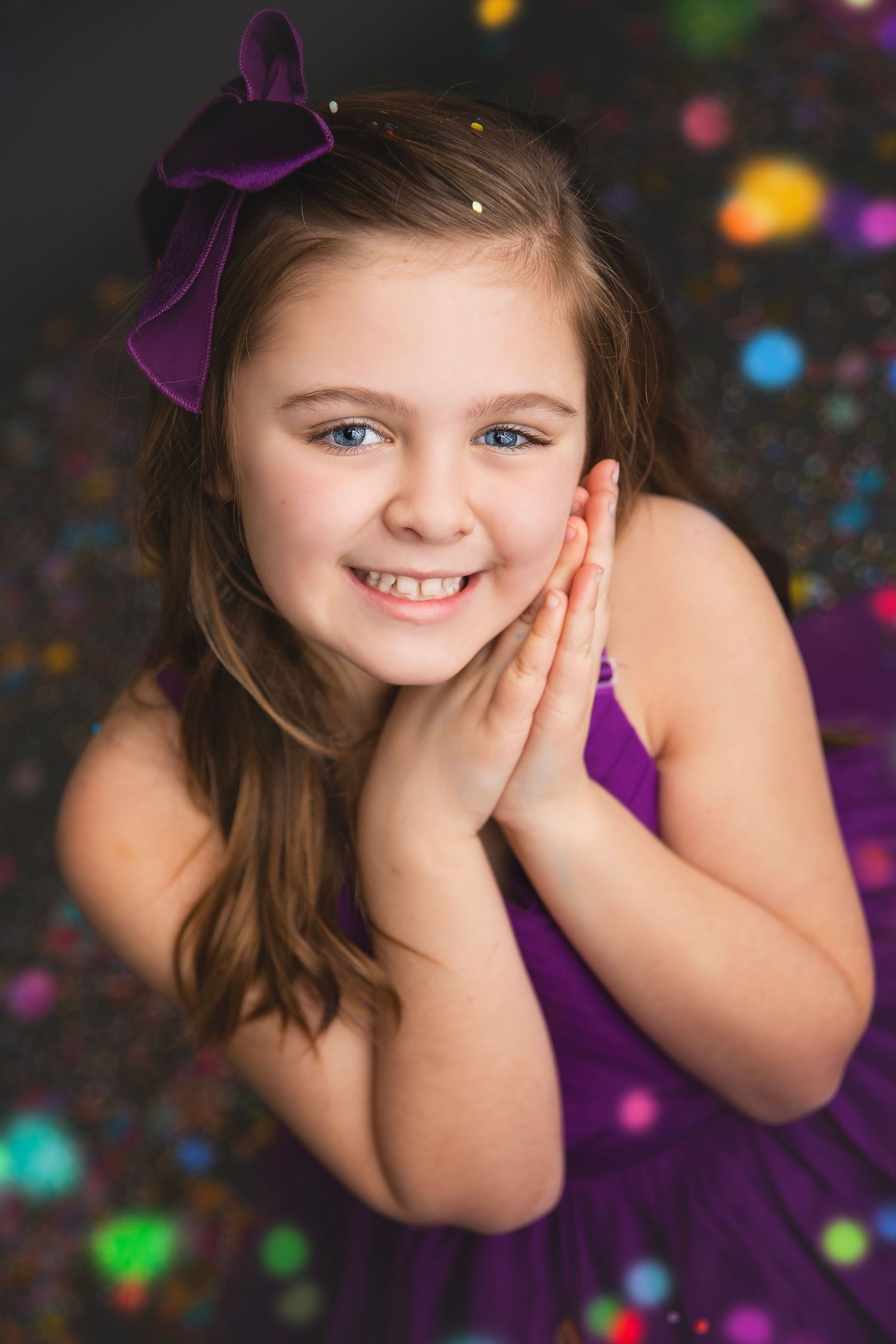 Smiling girl in purple dress with bow headband, hands on cheeks, surrounded by colorful bokeh lights.