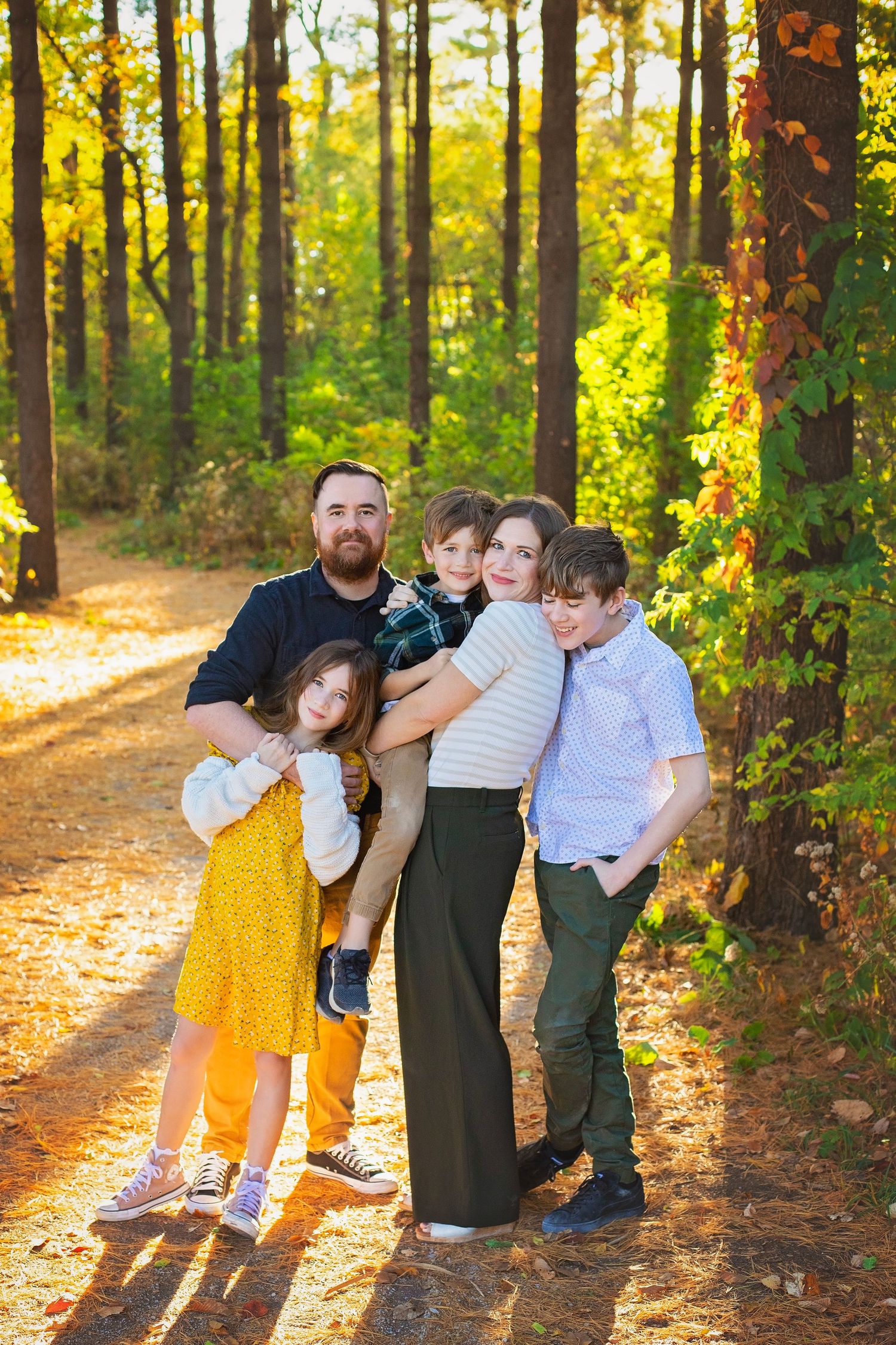 Family of five hugging on a golden sunlit path through an autumn forest.