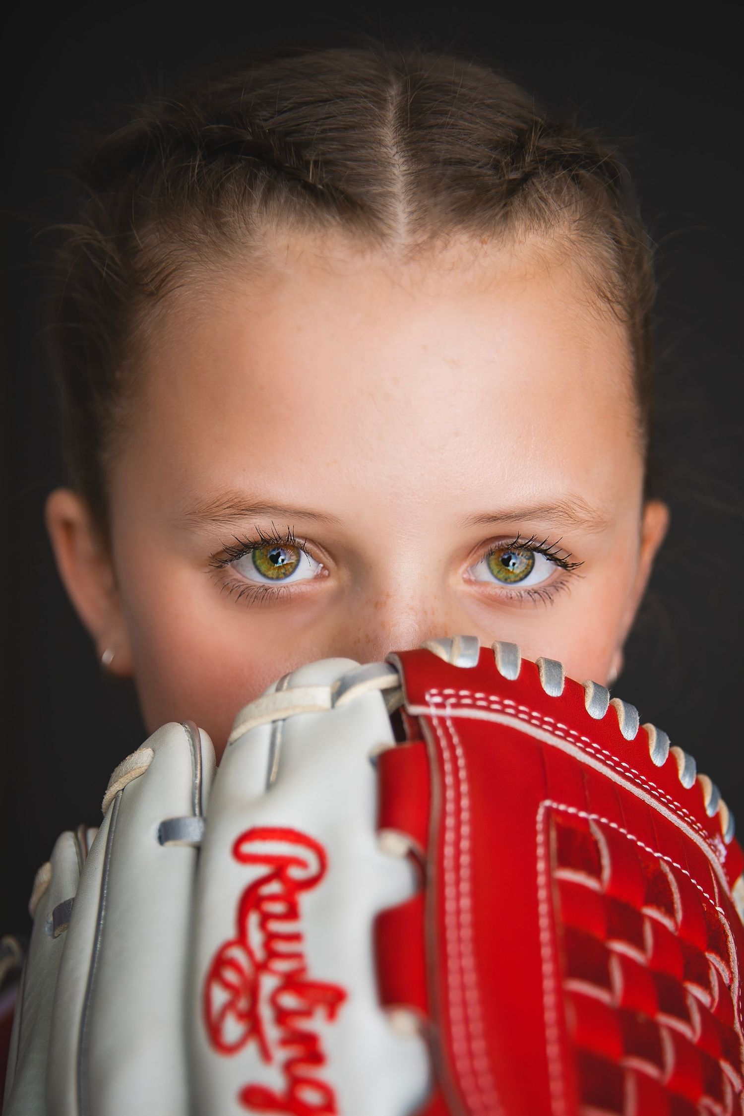 Close-up portrait of young softball player with striking eyes peering over red Rawlings glove.