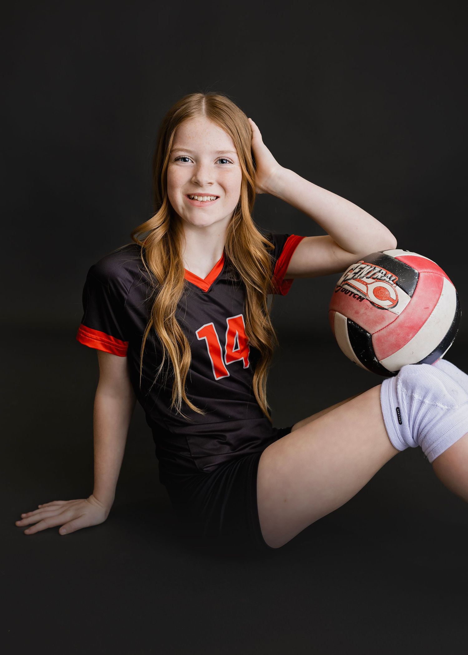 Smiling girl in number 14 volleyball jersey sitting on floor holding volleyball against dark background.